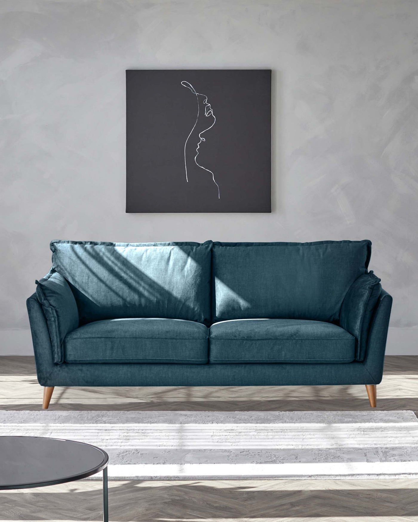 A modern teal blue fabric sofa with three seat cushions and cylindrical wooden legs, accompanied by a round glass-top coffee table with a thin black metal frame, placed on a white and grey patterned rug. A monochromatic artwork hangs above the sofa, adding a touch of elegance to the scene.