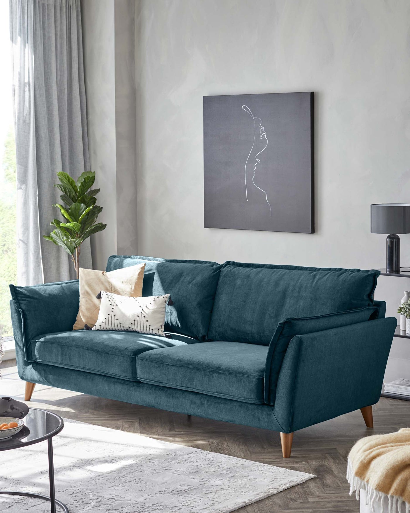Elegant three-seat sofa upholstered in a plush teal velvet, featuring a clean-lined silhouette with structured arms and back cushions, complemented by three decorative throw pillows in coordinating colours. The sofa stands on tapered wooden legs, set within a modern living room setting with neutral tones.