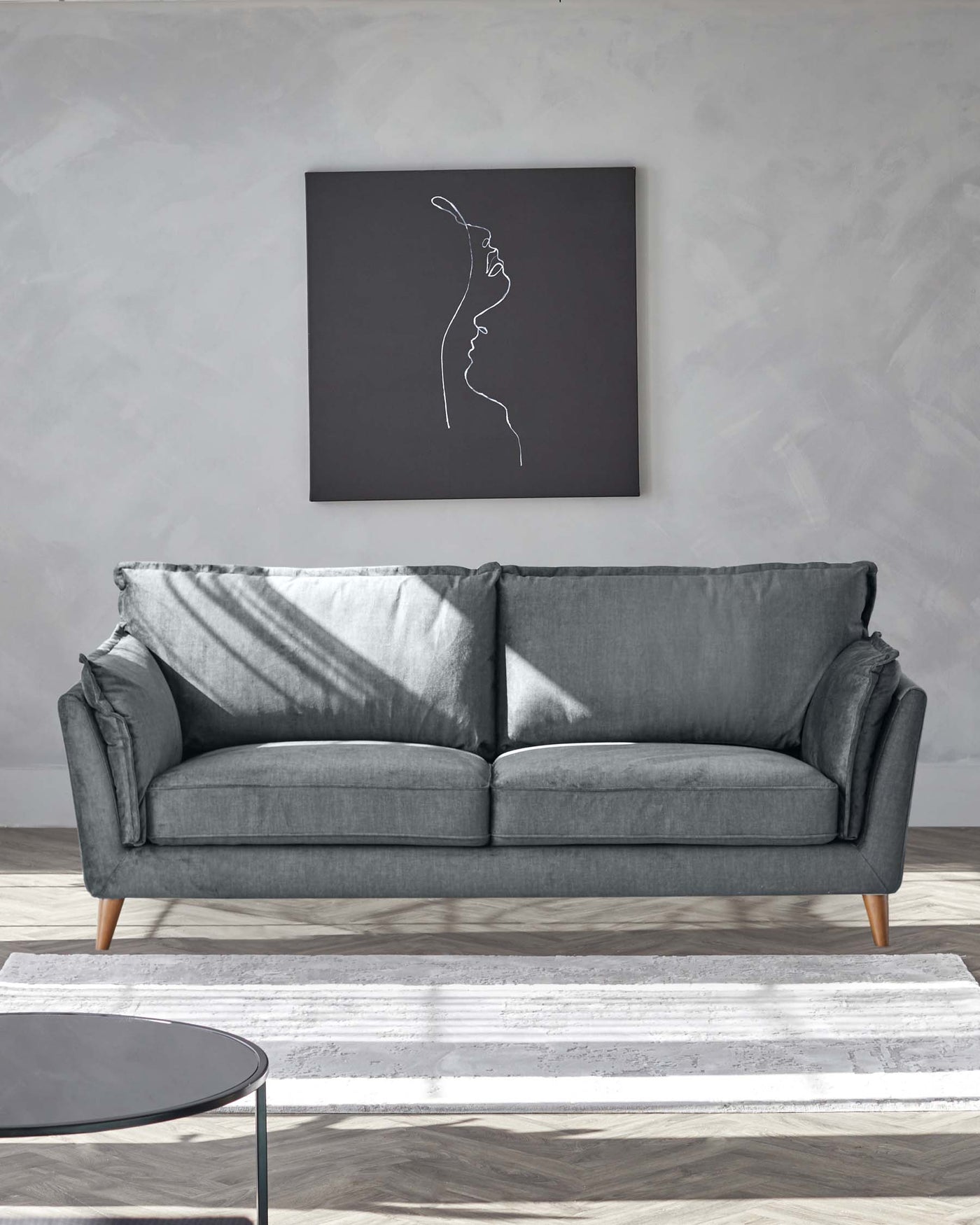 Modern grey fabric three-seater sofa with cushioned back and seat, flanked by tapered wooden legs. In front of the sofa is a round, black top coffee table with thin metal legs, complementing a white textured area rug underneath, creating a contemporary living space.
