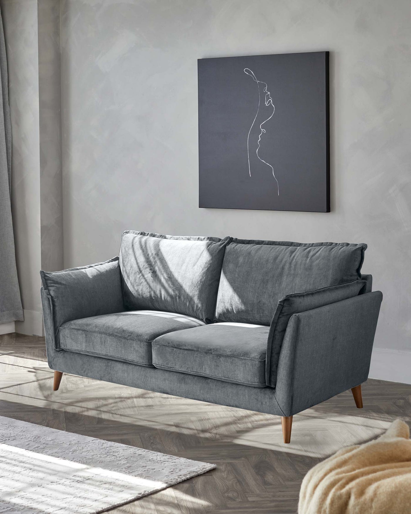 Elegant modern sofa in a soft grey fabric with three plush seat cushions and three back cushions, featuring square arms and angled wooden legs. The setting includes a light grey area rug on hardwood flooring, with a textured throw blanket partially visible in the foreground.
