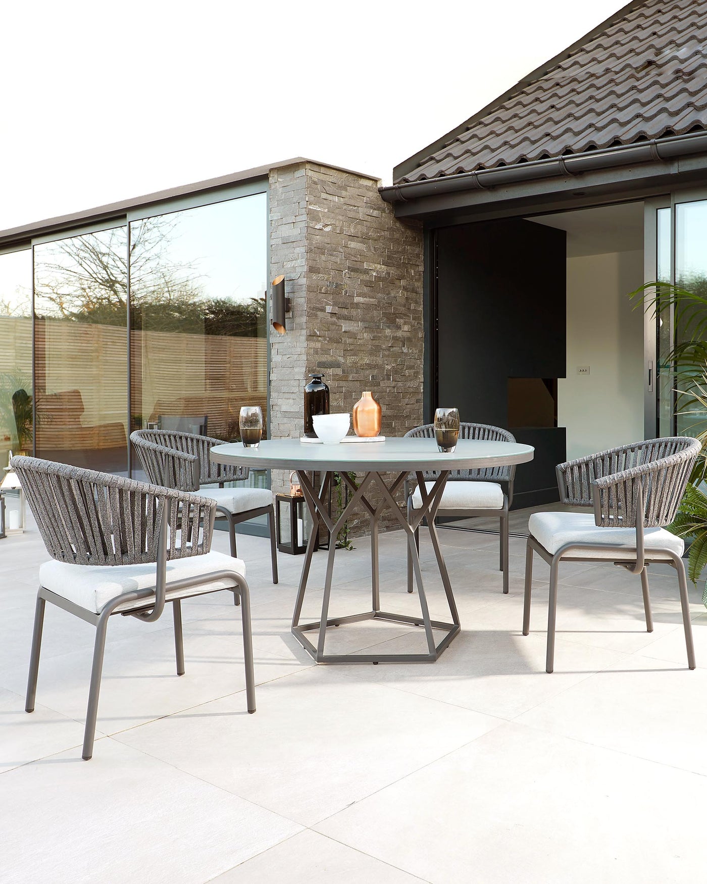 Contemporary outdoor dining set featuring a round, smoky glass-topped table with a unique geometric metal base, and four matching armchairs with woven backrests and seat cushions.