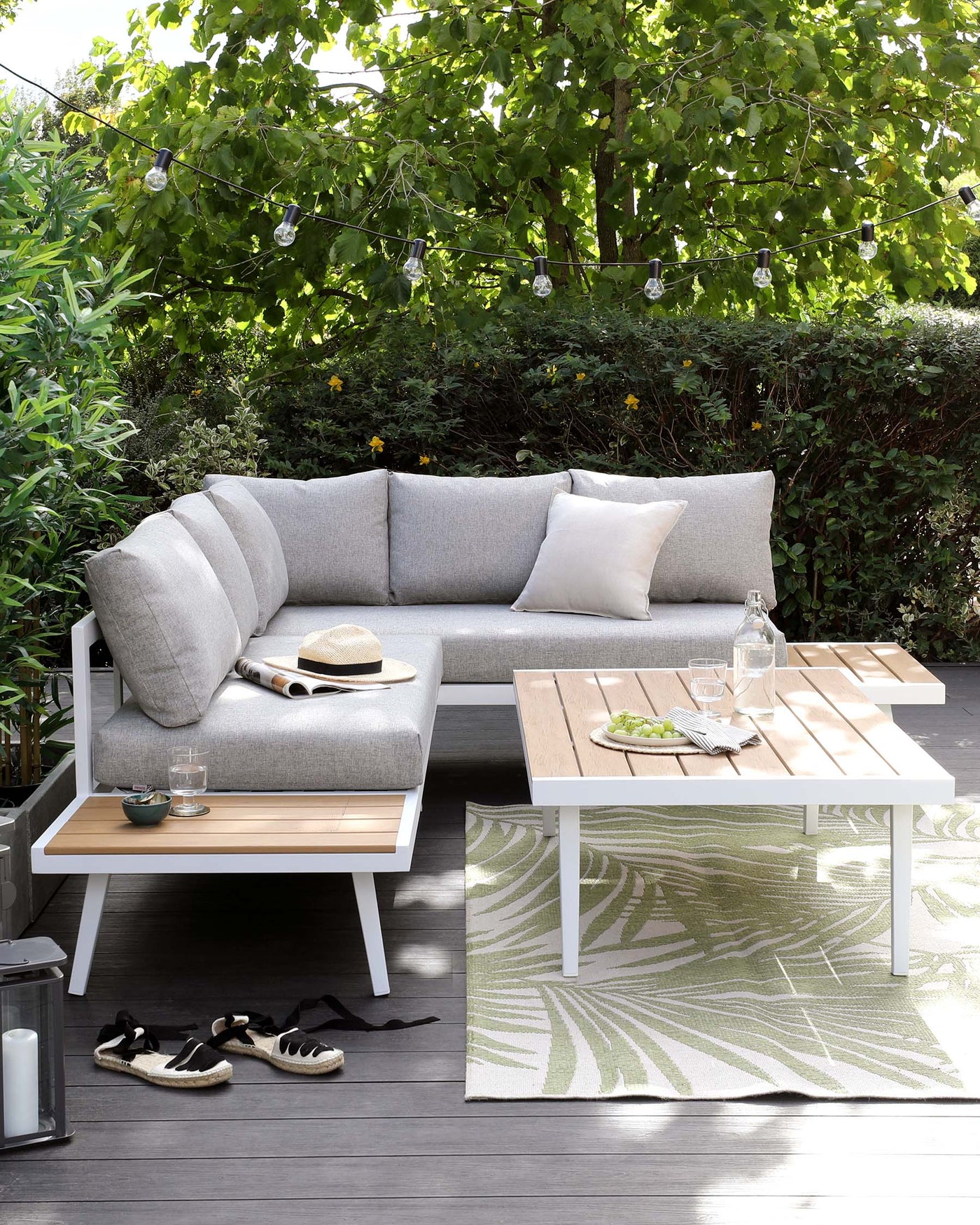 Outdoor furniture set including a light grey upholstered corner sectional sofa with matching cushions, a rectangular wooden coffee table with a white frame, and a matching side table with partially visible wooden slats on top. The set is complemented by a patterned area rug underneath the coffee table. The furniture arrangement is on a wooden deck, surrounded by lush greenery and adorned with string lights overhead.