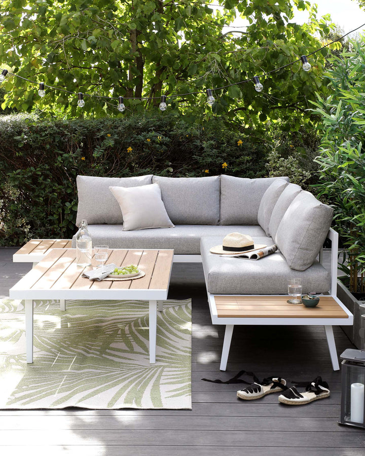 Outdoor contemporary furniture set featuring a modular corner sofa with light grey cushions and a white low-profile coffee table with a slatted wooden top. The set is arranged on a patterned area rug with leaf motif.