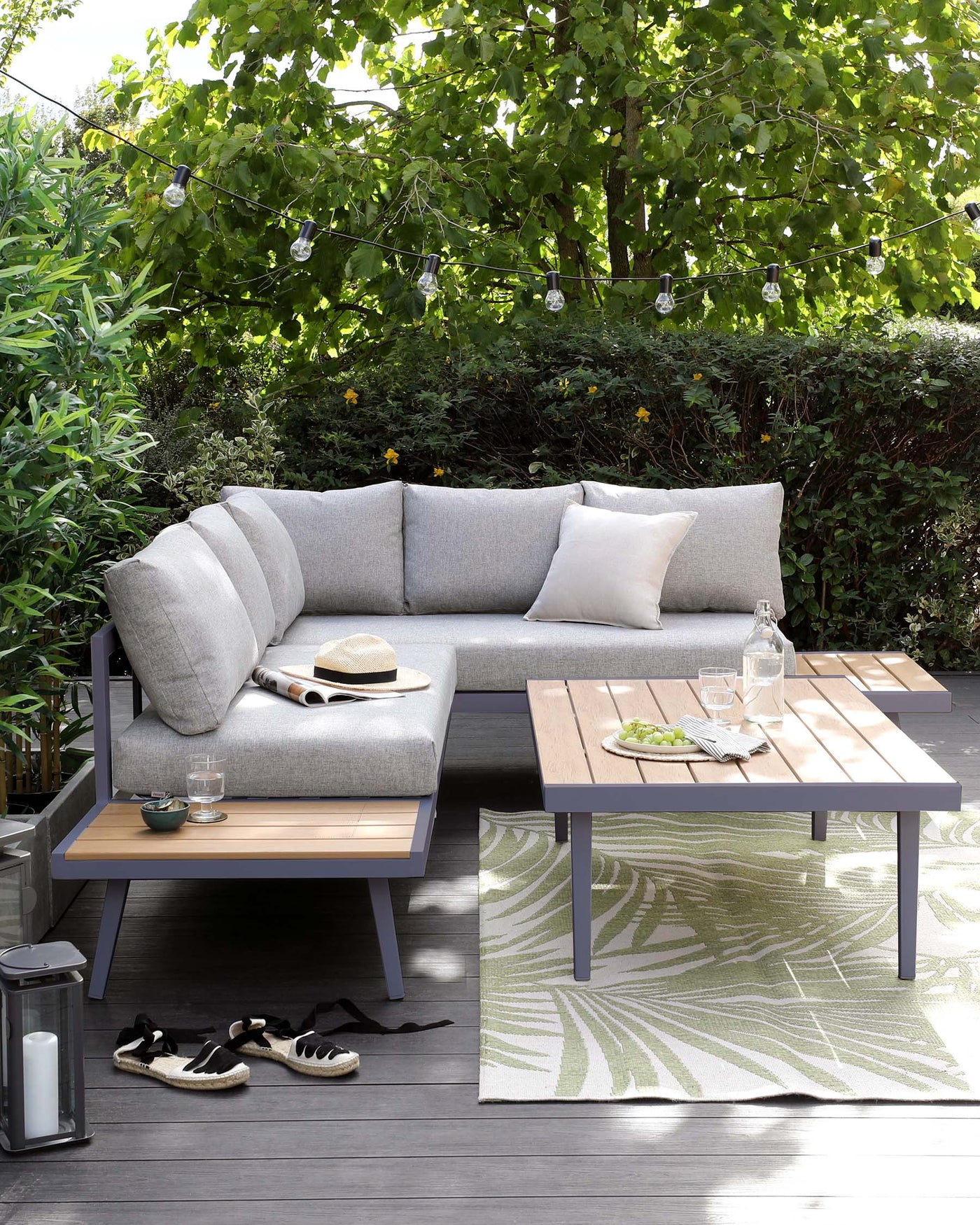 Modern outdoor furniture set featuring a grey upholstered corner sofa with additional cushions and a rectangular coffee table with a natural wood finish top and grey legs. Accessories such as a hat and sneakers give a lived-in look, all displayed on an outdoor rug with a leaf pattern, set against a backdrop of lush greenery and strung patio lights.