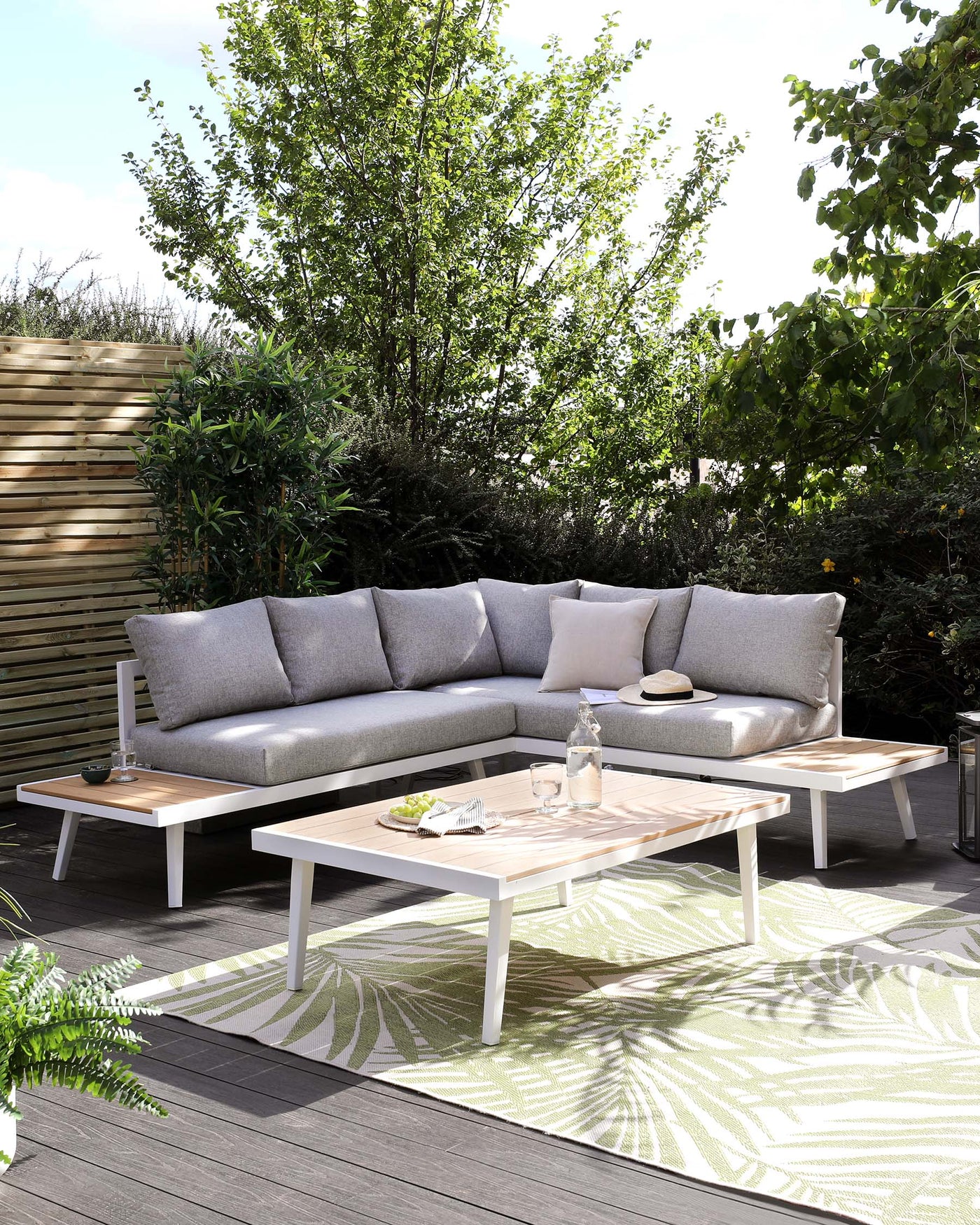 Outdoor sectional sofa with light grey cushions and a natural wood frame, accompanied by two matching coffee tables with white legs and a slatted wood top. The arrangement is placed on a deck with a patterned outdoor rug, enhancing the cosy ambiance.
