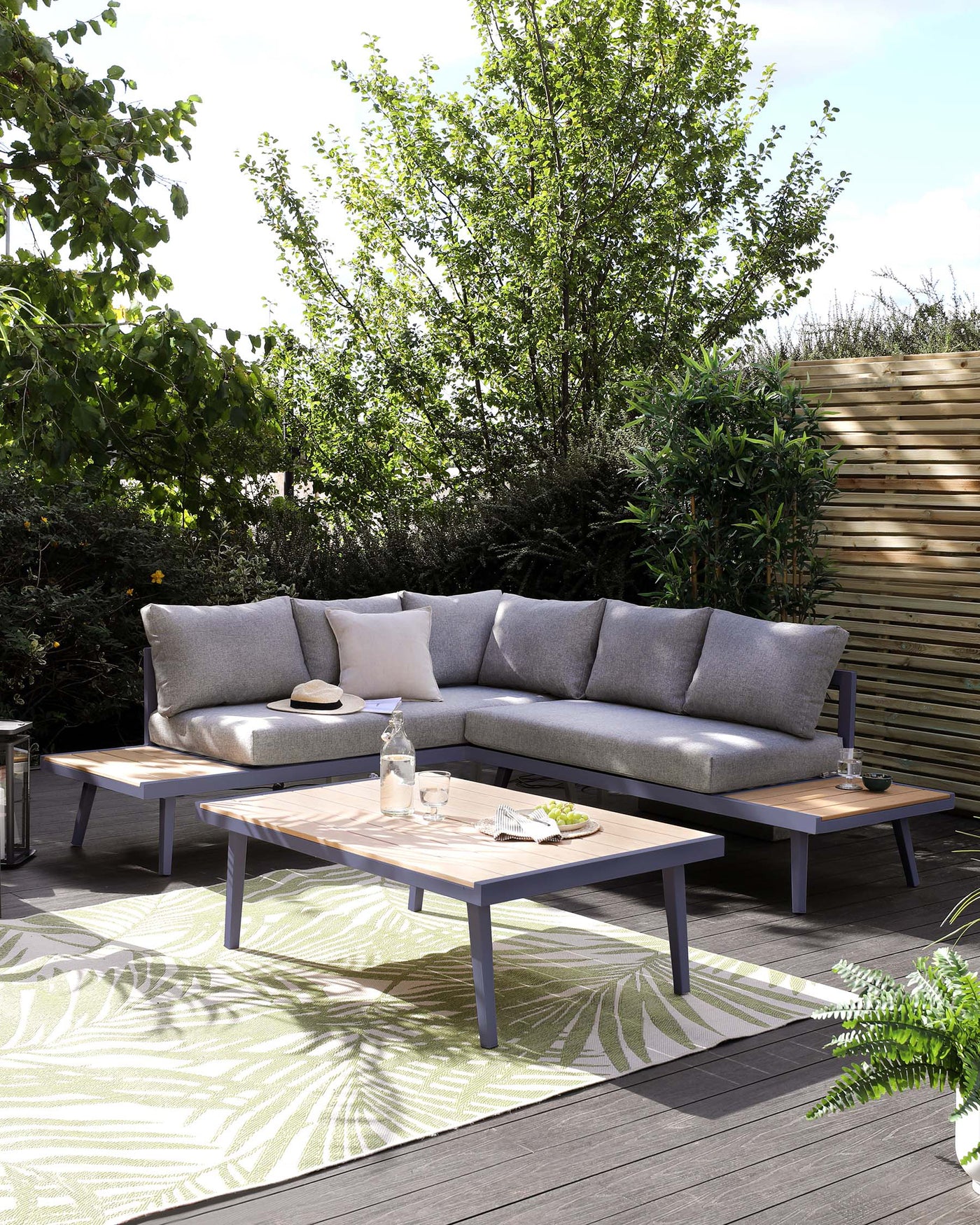 Modern outdoor furniture set including a sectional corner sofa with grey cushions on a wooden frame and two matching wooden tables with grey metal legs, displayed on a deck with a green patterned outdoor rug.