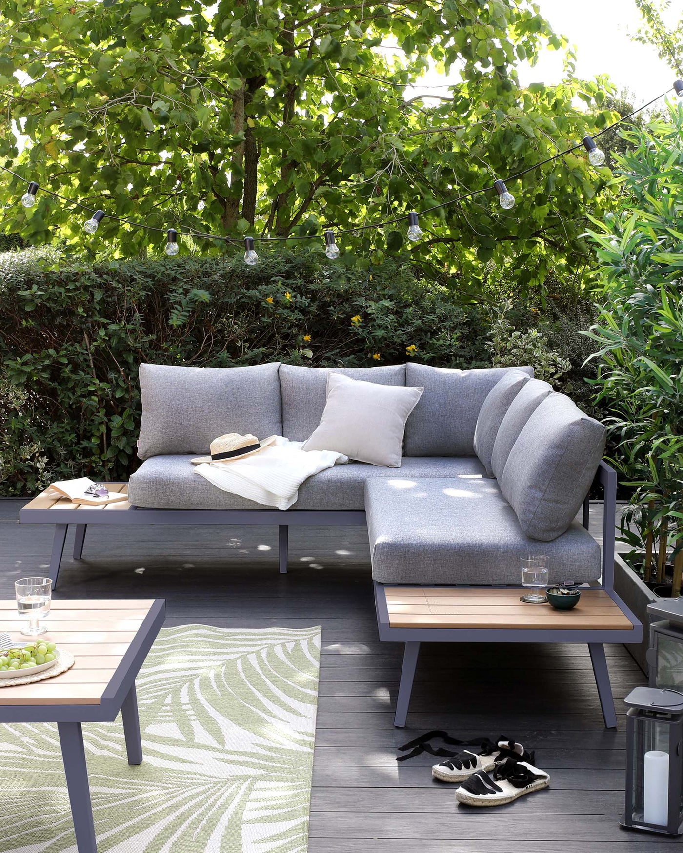 Modern outdoor furniture set featuring a grey sectional sofa with plush cushions and a matching low-profile coffee table with a wooden tabletop. The setting is completed by a patterned outdoor rug and atmospheric string lights overhead, all set against a lush backdrop of greenery and a wooden deck.