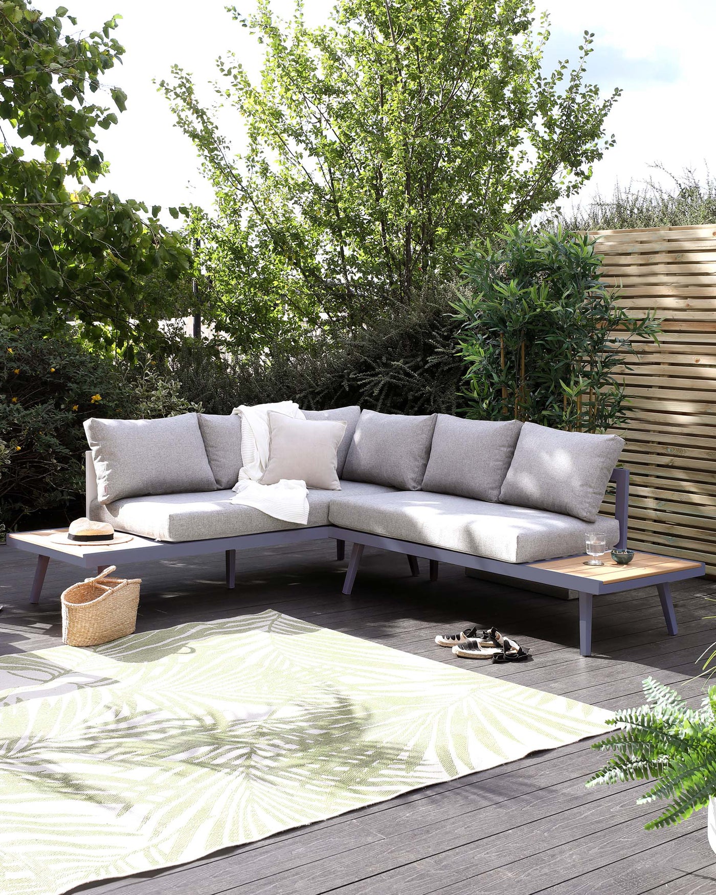 Outdoor sectional corner sofa with light grey cushions on a minimalist wooden frame, accompanied by a modern low coffee table with a dark grey top and light wooden legs, set upon an ornate green and white patterned area rug. A woven wicker basket adds a touch of natural texture to the setting.