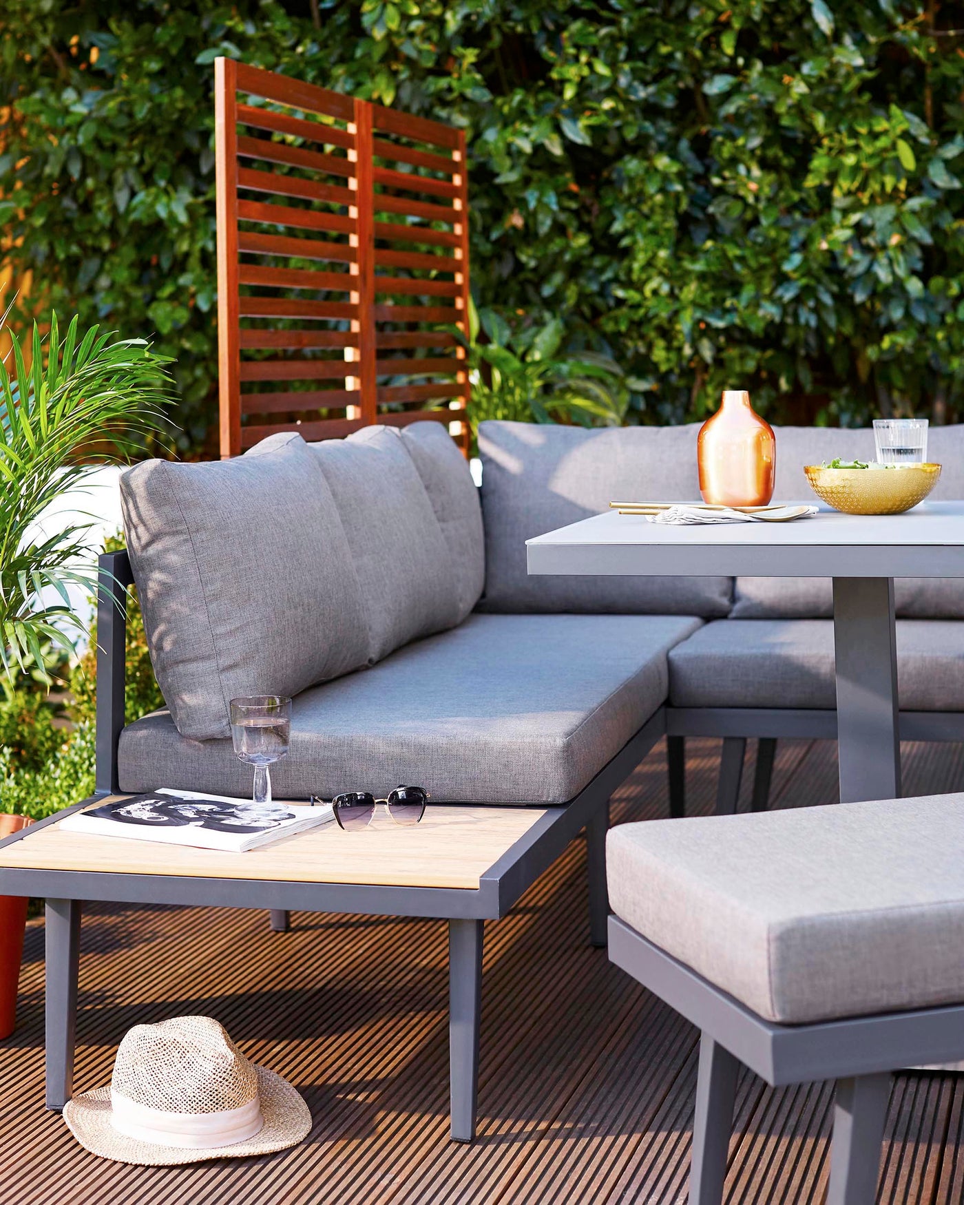 Outdoor patio setup featuring a modern modular corner sofa with light grey cushions, a square table with a natural wood top and grey metal frame, and a minimalist wooden slatted screen in the background, all set against lush green foliage.