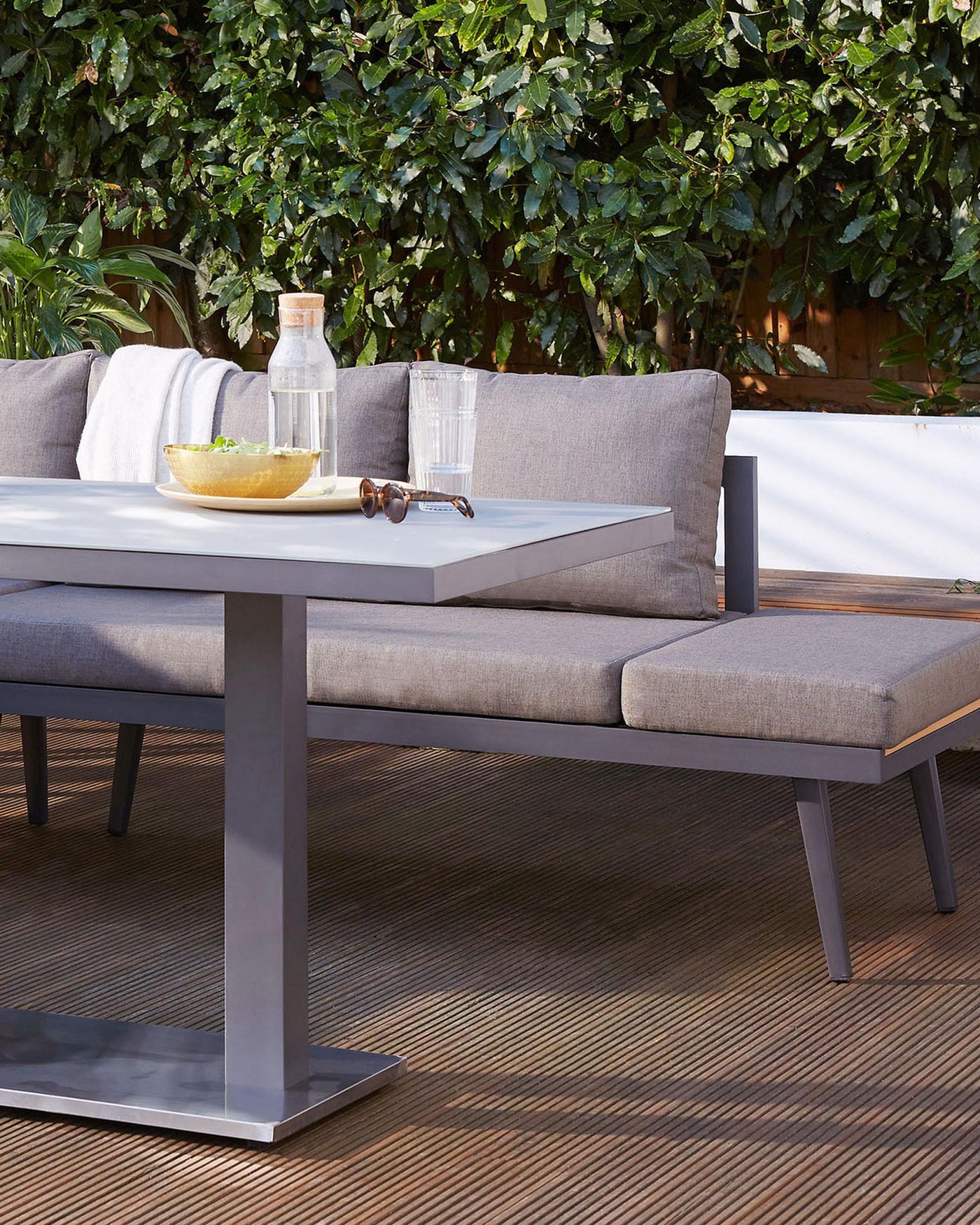 Modern outdoor furniture set featuring a sleek, grey aluminium frame. Includes a rectangular low-profile coffee table with a white tabletop and a matching corner sectional sofa with light grey cushions and pillows. The ensemble is complemented by a coordinating ottoman resting against the sectional. Set against a backdrop of lush green foliage, creating a serene outdoor living space ambiance.