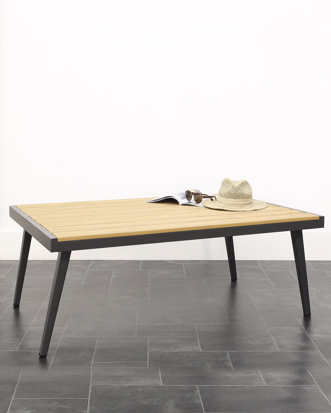 A modern rectangular coffee table with a ribbed light wooden tabletop and slender, straight metal legs in a matte black finish. The table is adorned with a casual straw hat and a pair of sunglasses resting atop an open magazine. The furniture piece is set against a neutral background with grey floor tiles.