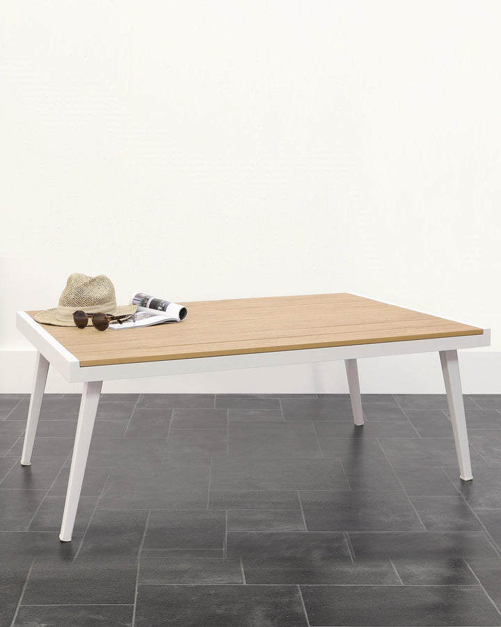 Modern rectangular coffee table with a light wooden tabletop and white legs, set against a plain white wall and dark tiled flooring. Accessories including a woven hat, sunglasses, and an open magazine are placed on the tabletop.