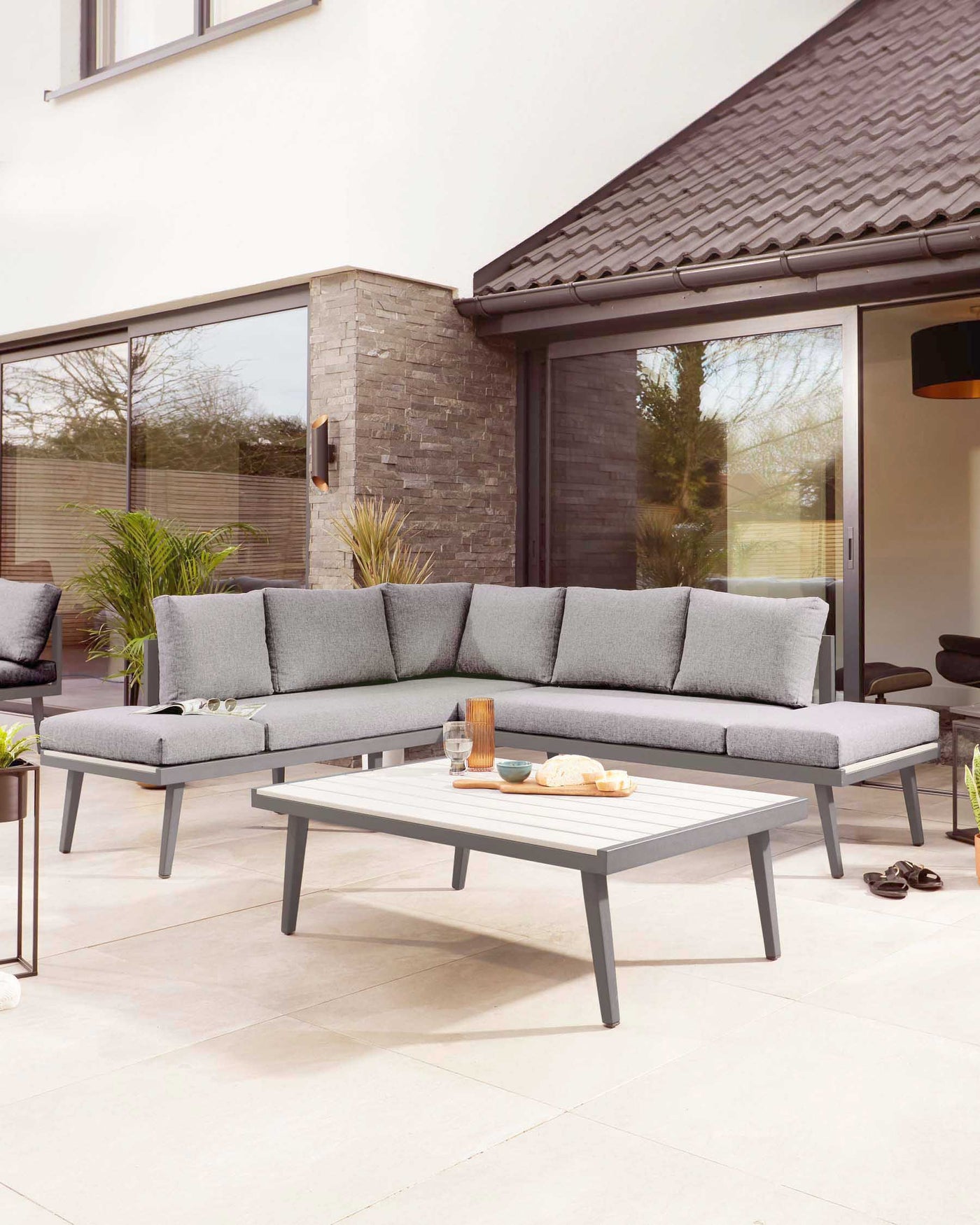 Modern outdoor lounge set featuring a sectional sofa with grey cushions and a sleek, grey aluminium frame, paired with a rectangular coffee table with a white tabletop and matching grey frame, all displayed on a patio setting.