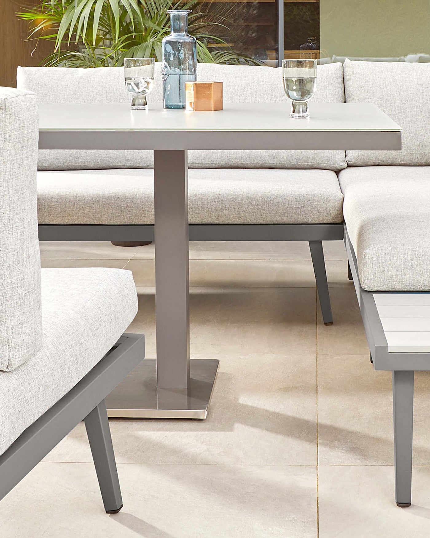 Modern outdoor furniture featuring a minimalist light grey rectangular dining table with a sturdy central metal pedestal base and a sleek matching bench with light taupe cushions. The set projects a contemporary, understated style suitable for urban terraces or chic garden spaces.
