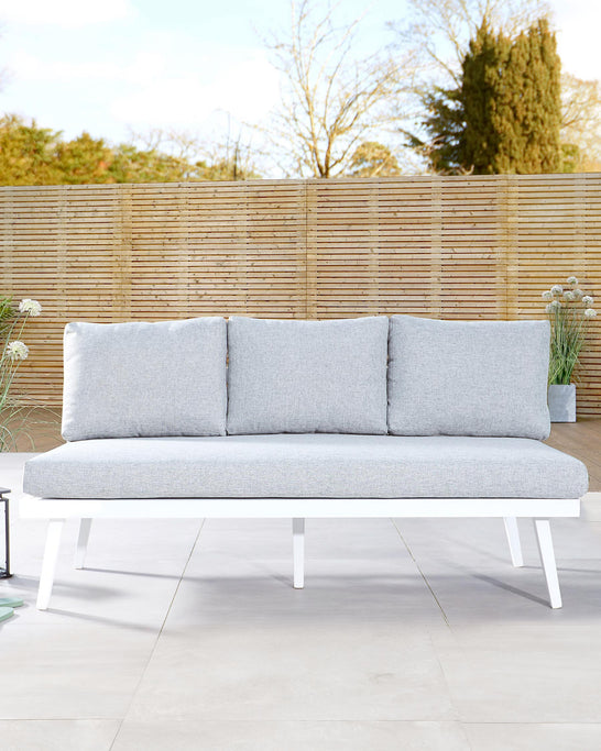 Modern outdoor three-seater sofa with a minimalist white frame and light grey cushions, displayed in an outdoor setting with a wooden fence and greenery in the background.
