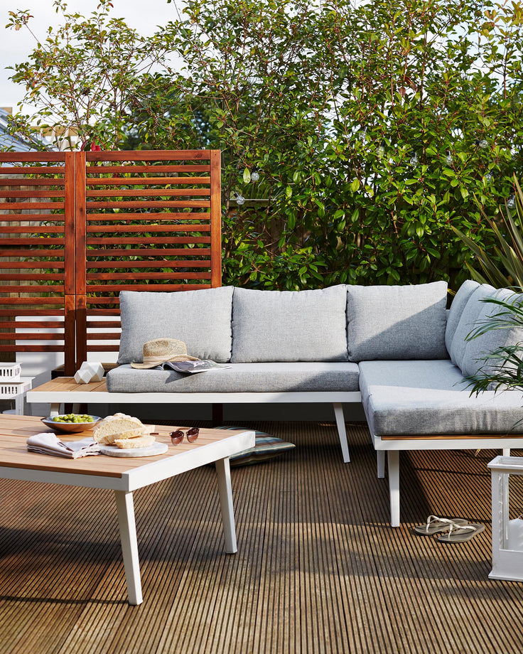 Modern outdoor furniture set featuring a sectional sofa with light grey cushions, a white metal frame, and teak wood accents. A matching low white coffee table with a teak wood top sits in front, adorned with a casual spread of food and a magazine, creating an inviting terrace or patio setting.