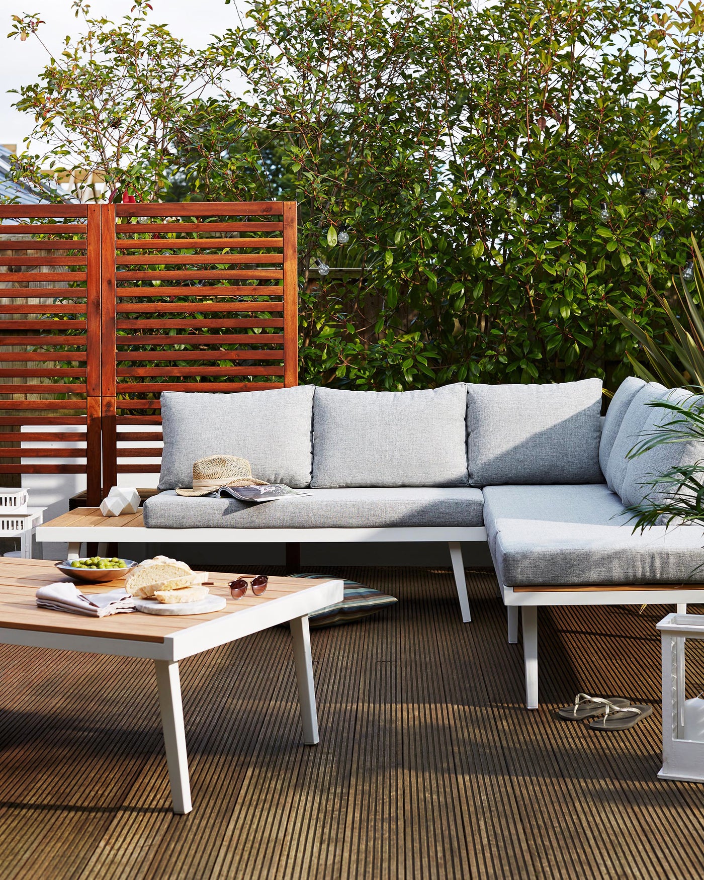 Outdoor corner sofa set with light grey cushions, a white and wooden rectangular coffee table, and two white side stools with a slatted design set on a deck with lush greenery in the background.