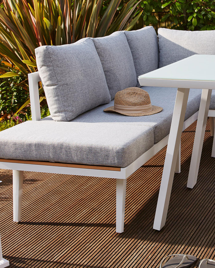 Modern outdoor furniture set featuring a white-framed corner sofa with light grey cushions and a coordinating white square table, displayed on a warm brown deck with lush greenery in the background. A straw hat rests on the sofa, adding a relaxed, summery vibe.