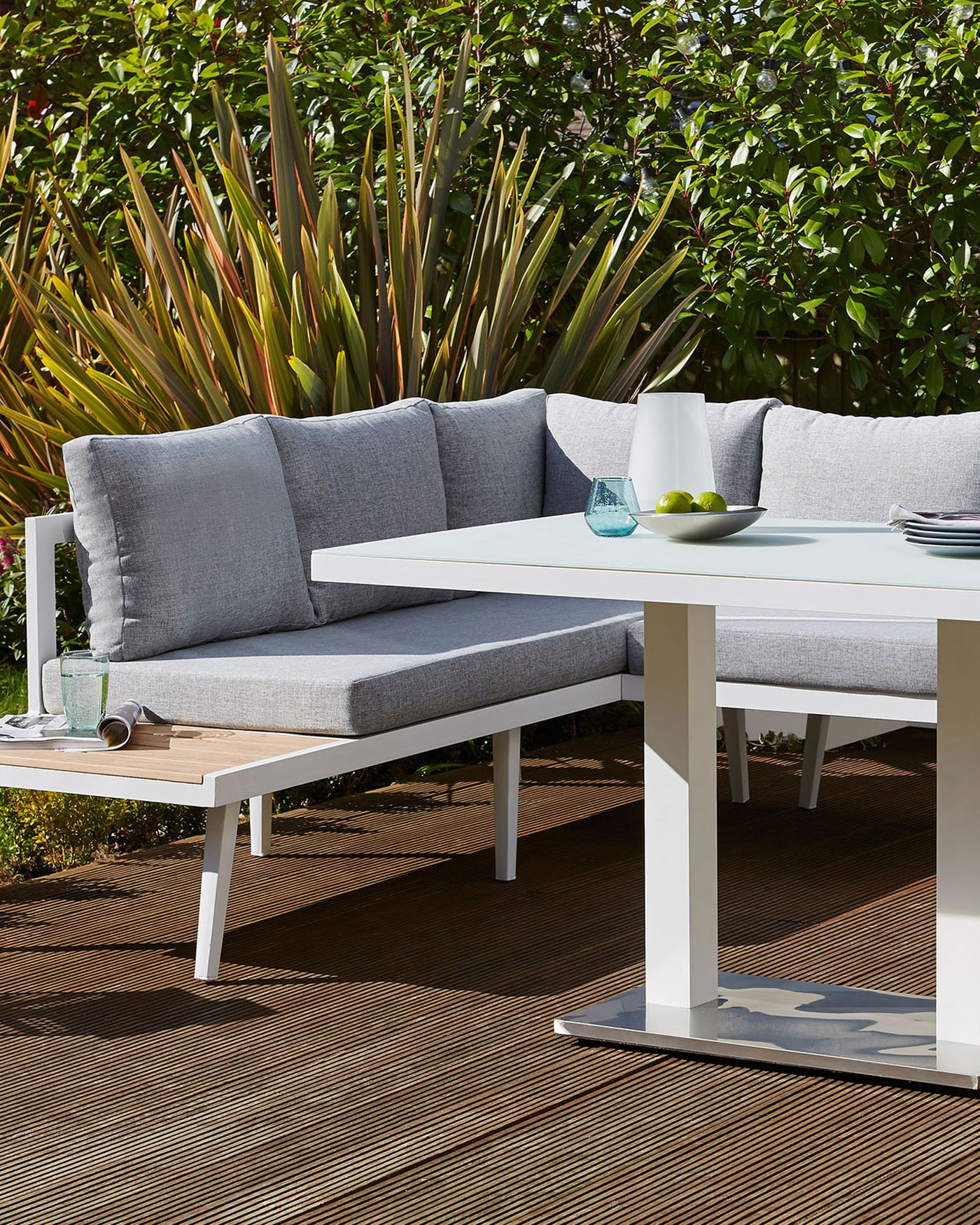 Contemporary outdoor furniture set featuring a corner sectional sofa with light grey cushions and a matching white rectangular coffee table, displayed on a wooden deck with lush greenery in the background.