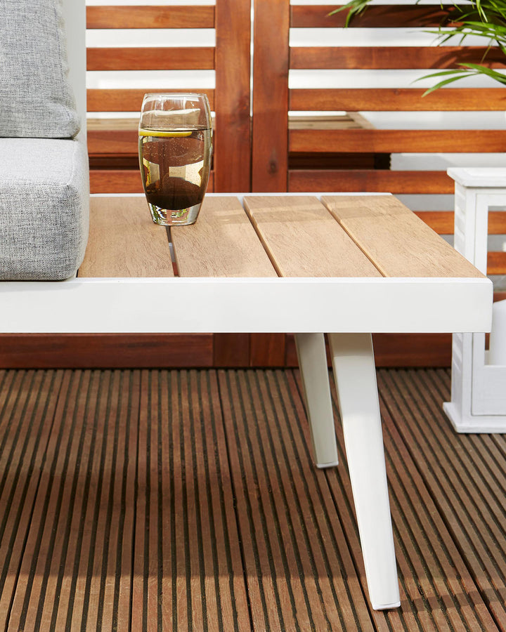 Modern outdoor furniture set featuring a sleek white-framed side table with a natural wood tabletop and a coordinating white-framed armchair with grey cushions visible on the left, set against a backdrop of a wooden patio and greenery.