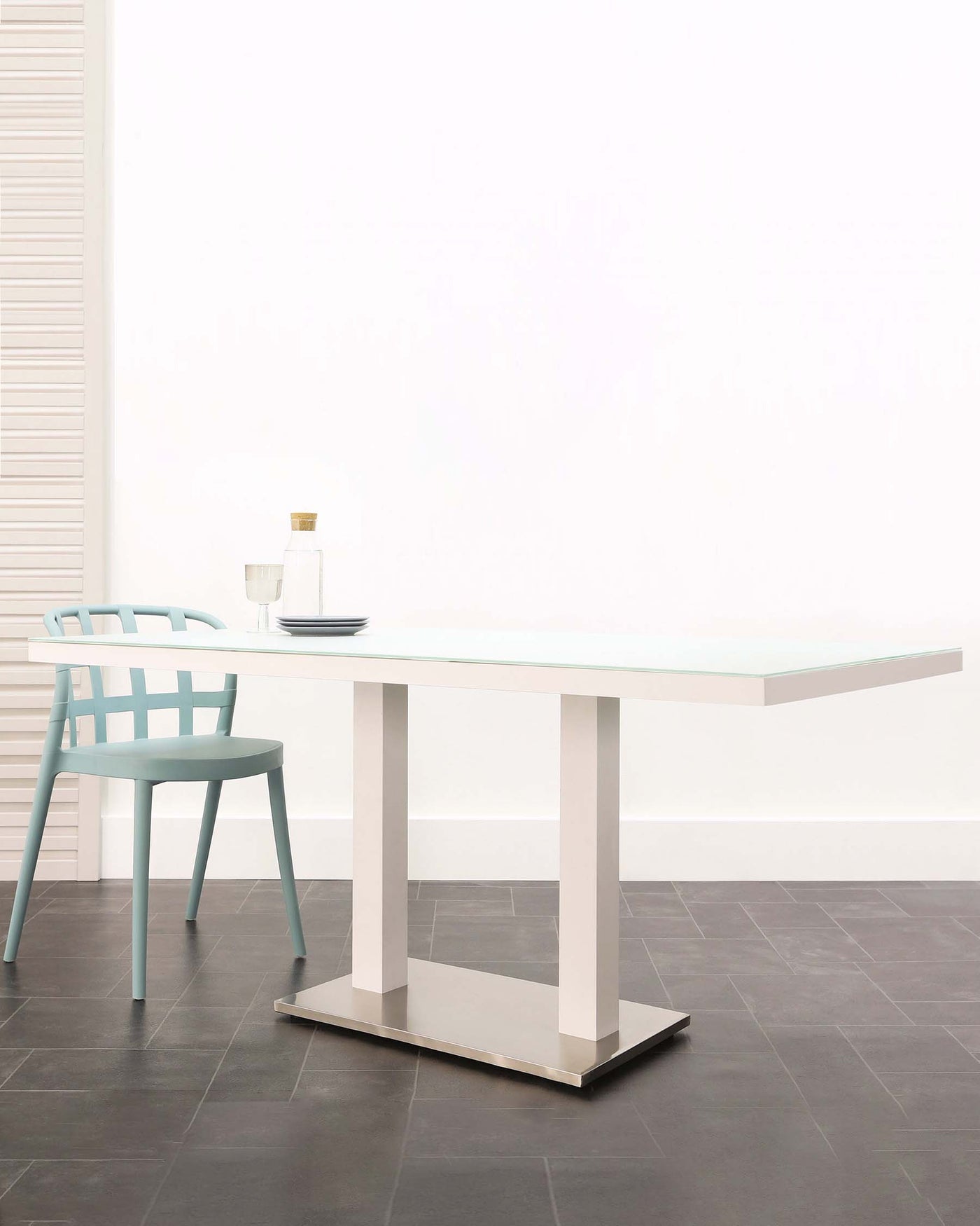 Modern minimalist furniture set featuring a white rectangular dining table with a sleek tabletop supported by two wide, flat legs connected at the base, paired with a single aqua blue chair with a curved backrest and slender legs. The set is displayed in a bright room with light walls and dark floor tiles, accented by a simple decorative bottle and glass placed on the table.