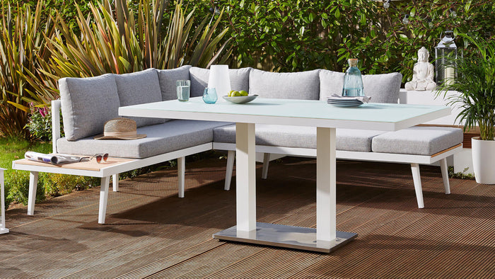 Outdoor garden furniture set featuring a modern white rectangular table with a light green frosted glass top and sturdy white frame, accompanied by a matching white corner bench with comfortable grey cushions, situated on a wooden deck with lush greenery in the background.