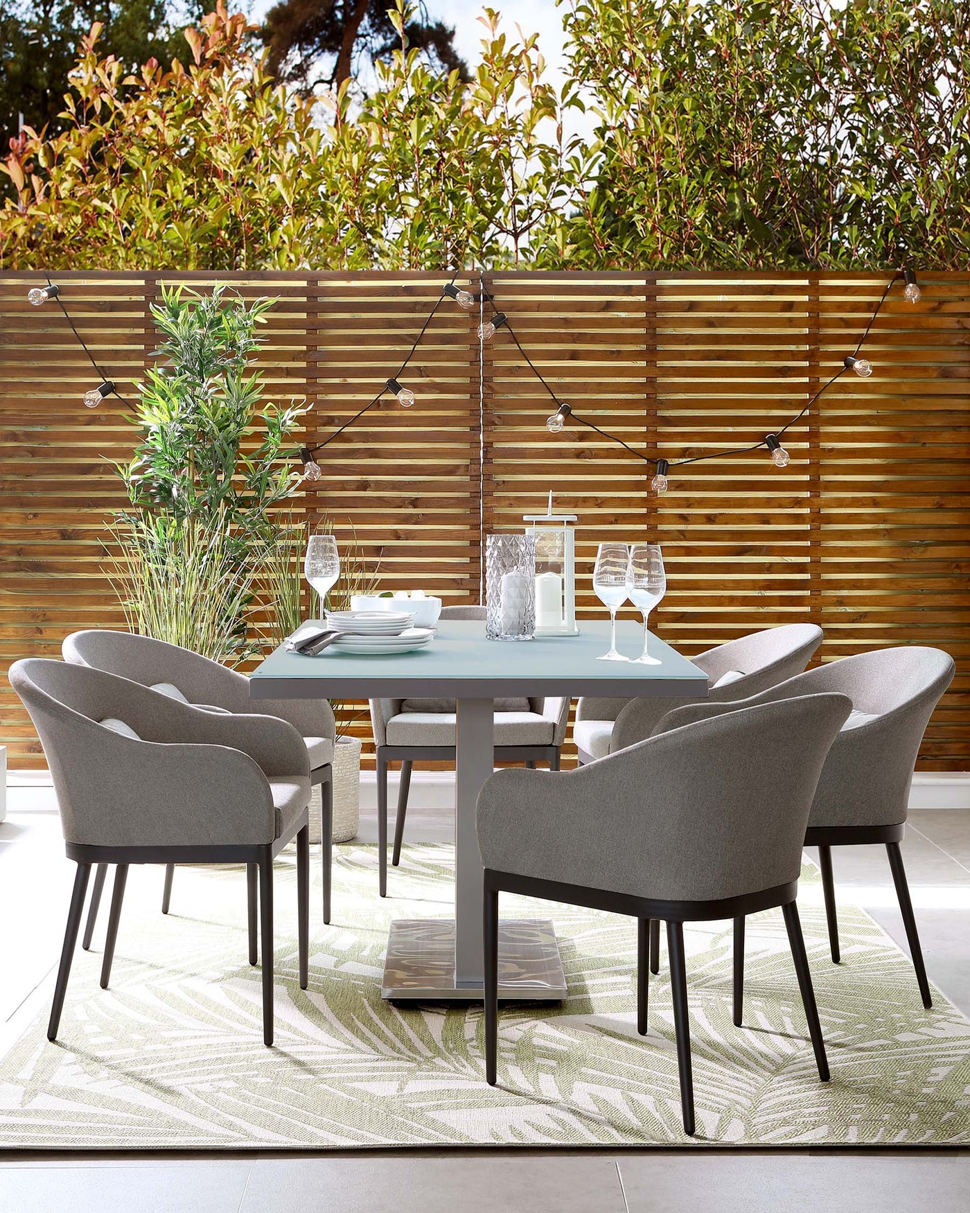 Elegant outdoor dining set featuring a round, light blue table with a sleek, grey central support, surrounded by six grey upholstered armchairs with wooden legs, all positioned on a leaf-patterned area rug.