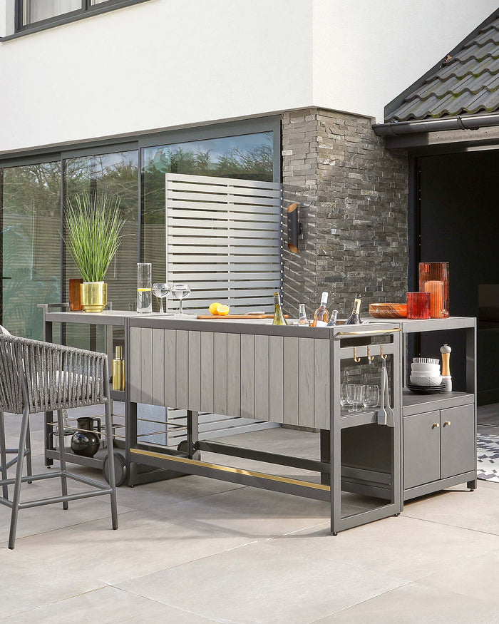 Outdoor modern grey modular kitchen island with black metal frame, featuring a built-in sink, storage shelves, and cabinets. Includes high stools with grey woven backrests and black metal legs. Accessories on the countertop include gold and coloured glassware, bottles, and a potted plant. The setup is on a patio with large glass doors leading to a home interior.