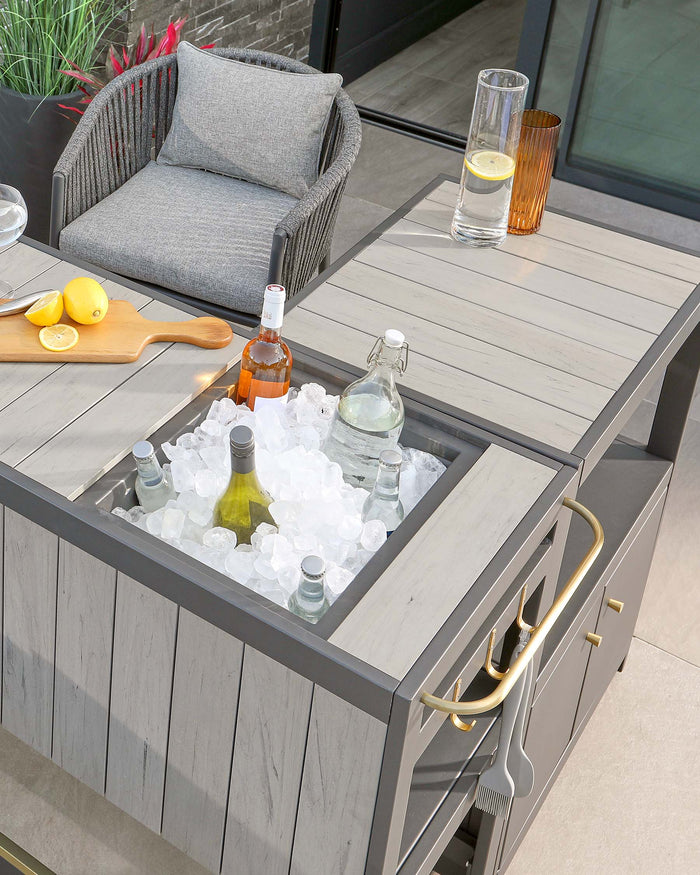 Modern outdoor furniture featuring a grey woven lounge chair with a plush cushion and a multifunctional grey wooden table with a built-in ice bucket for beverage cooling, accented with brass-coloured handles and a white tabletop finish.