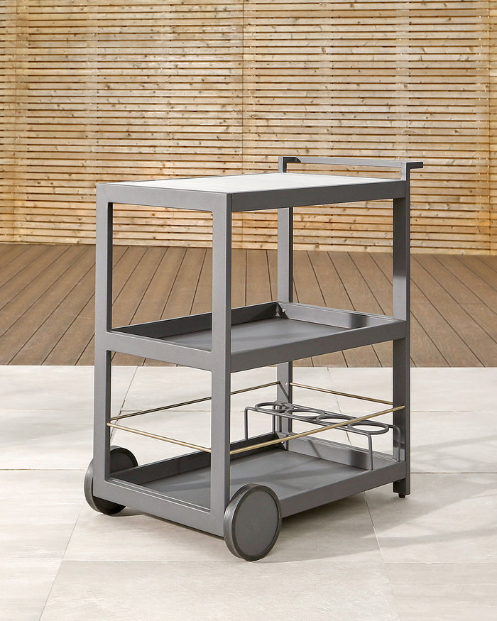 A modern grey rolling bar cart with three shelves, featuring a sturdy frame, a handle, and large round wheels. The top shelf includes a recessed tray area, while the middle shelf sports bottle holders, and the bottom shelf offers open storage space. The cart is showcased against a backdrop with a bamboo wall and wooden floor.