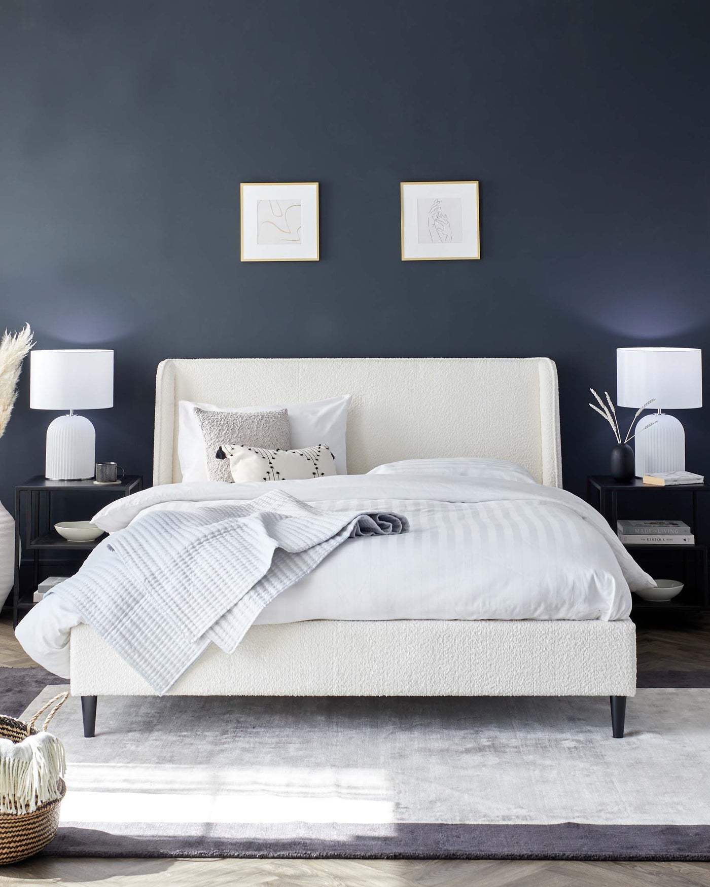 Elegant bedroom furniture set including an upholstered queen-sized bed in a creamy-white fabric with a high headboard and dark tapered legs. Accompanying the bed are two matching nightstands in black, featuring minimalist design, each with two drawers topped with white cylindrical table lamps and decorative items. A textured grey area rug lies beneath the bed, adding warmth and contrast to the light wooden flooring.