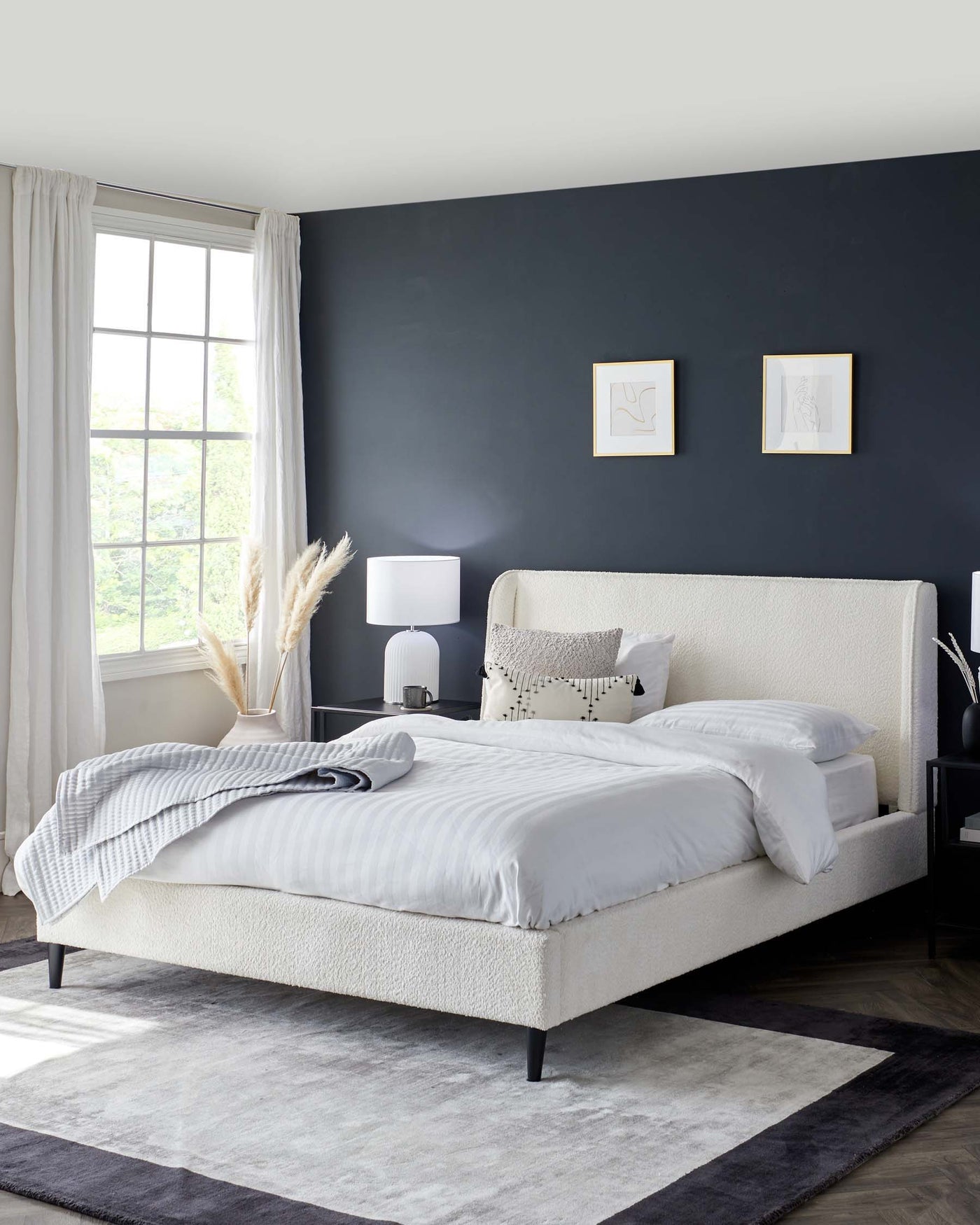 Elegant bedroom featuring a plush, upholstered cream bed frame with a high headboard, accompanied by two sleek black nightstands with clean lines and white table lamps. The bed is dressed in white bedding with minimalist pillows. A large grey and black area rug underlays the bed and nightstands, complementing the modern aesthetic of the room.