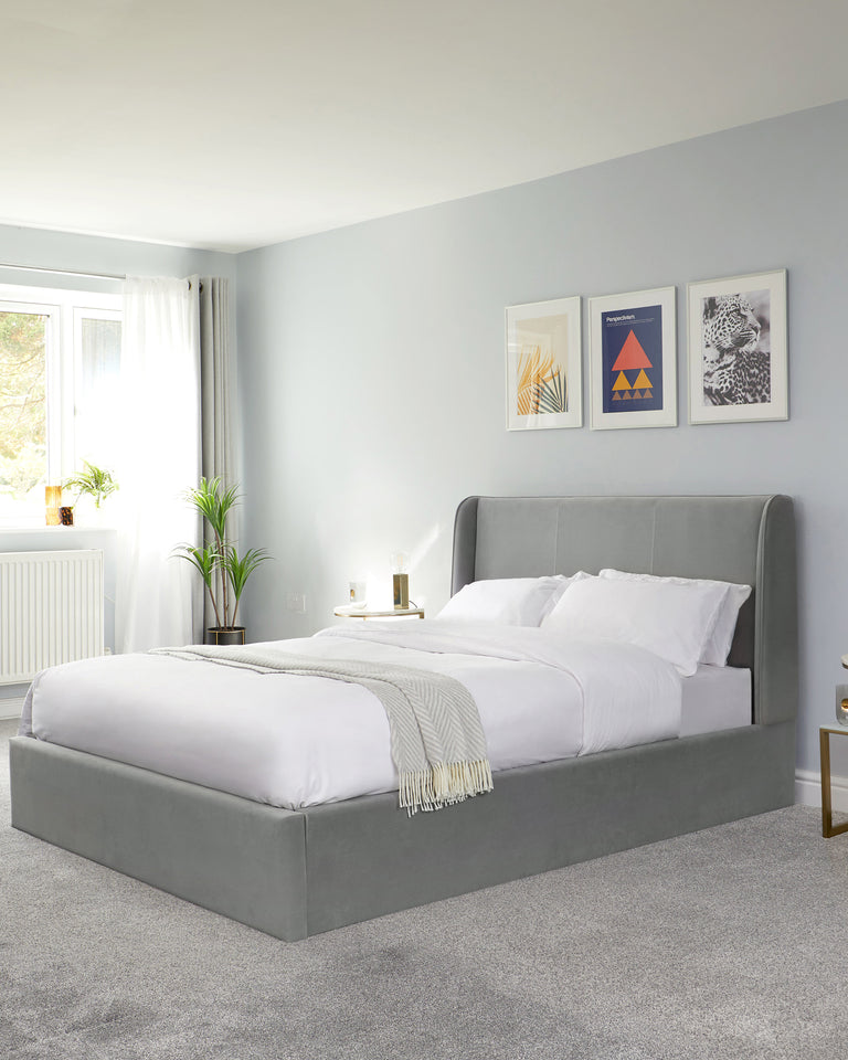 Contemporary grey upholstered platform bed with a high headboard in a room with white bedding and a decorative cream throw blanket. A side table with gold accents is visible to the right of the bed.