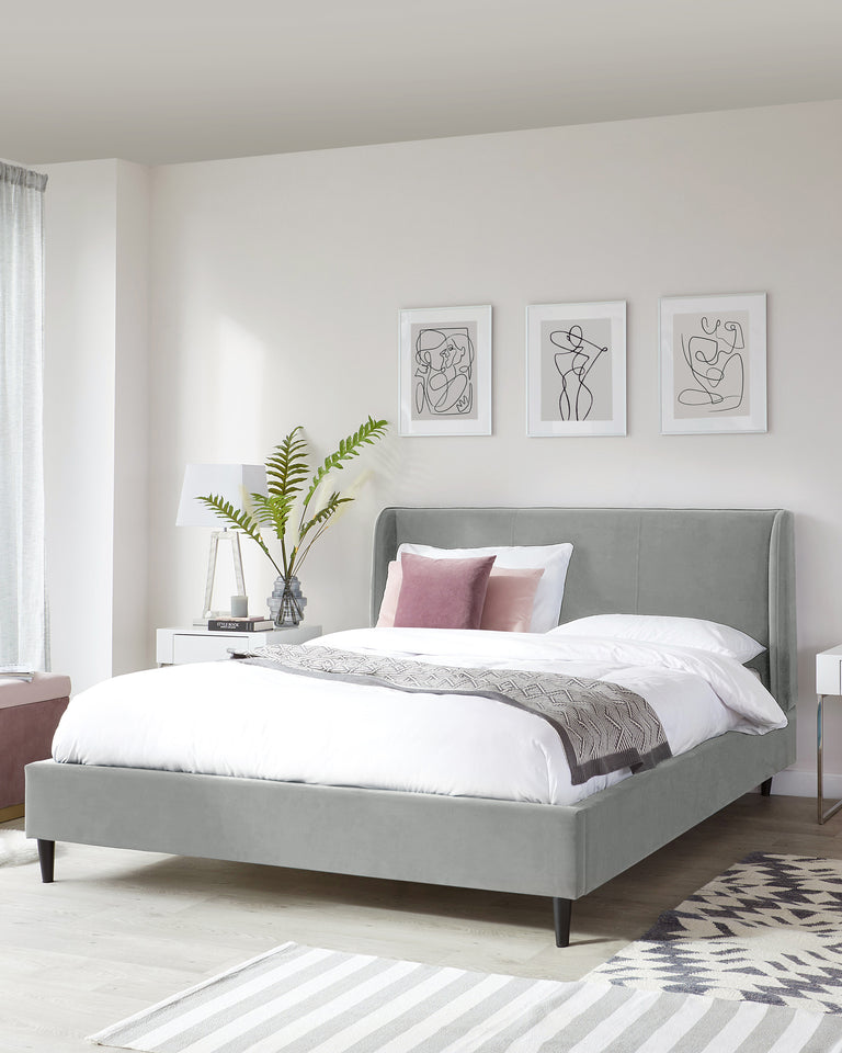 Modern king-sized platform bed with a wingback headboard upholstered in light grey fabric, featuring clean lines and dark stained wooden legs. A small sleek bedside table with a mirrored surface and a contemporary table lamp to the left. The bed is dressed with a white duvet, decorative grey patterned throw, and accent pillows in coordinating colours. An off-white and grey striped area rug is partially visible on the light wooden floor.