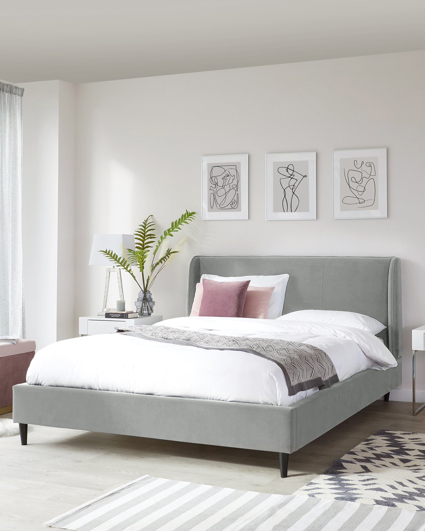 A modern upholstered queen-size platform bed in a light grey fabric with a high cushioned headboard, accompanied by a simple white nightstand with a glass vase and a table lamp, set against a neutral bedroom backdrop with minimalist decor.