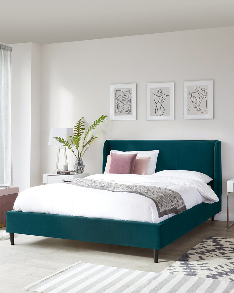 Elegant emerald green upholstered bed with a high headboard and dark wooden legs, flanked by a white modern nightstand with a lamp and decorative plant. A grey and white striped area rug lies at the foot of the bed.