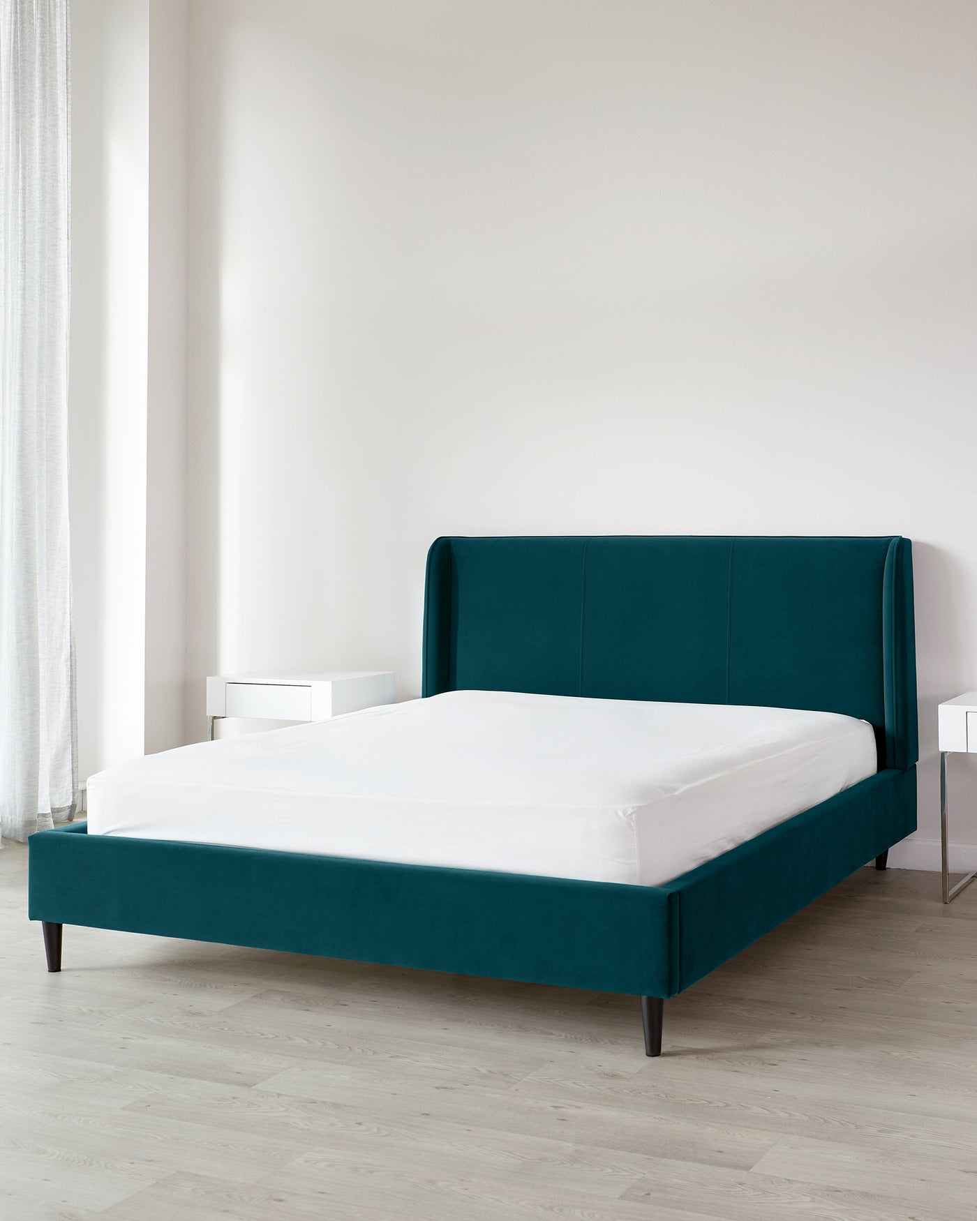 A contemporary emerald green upholstered bed frame with a high headboard, matched with two minimalist white bedside tables, showcased in a room with light wooden flooring and a sheer curtain.