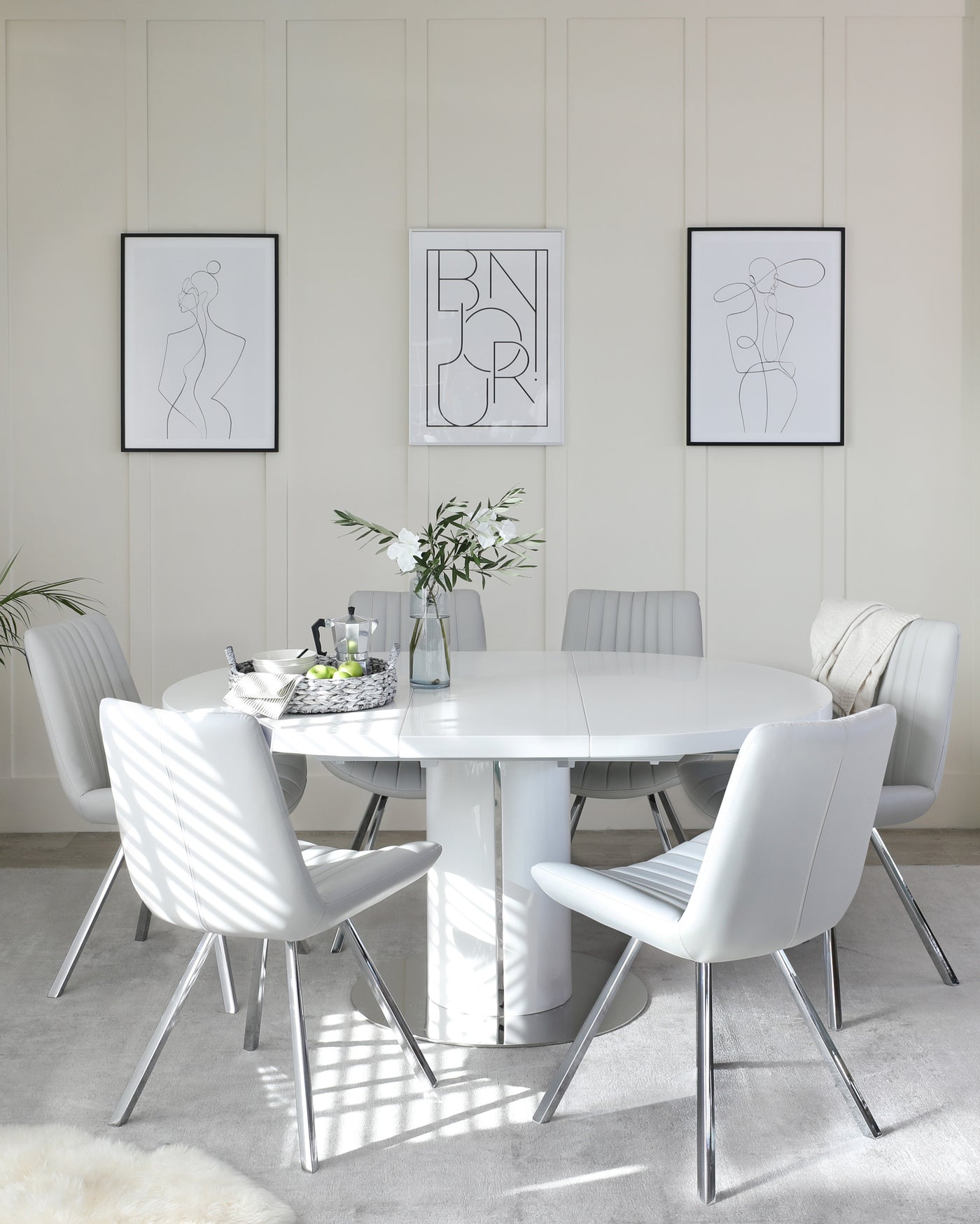 Modern dining room featuring a sleek, round white table with a glossy finish and a cylindrical base, surrounded by six contemporary light grey upholstered chairs with stylish chrome legs. The chairs display a horizontal channel stitch detail. A plush white area rug underlies the set, enhancing the clean, minimalist aesthetic.