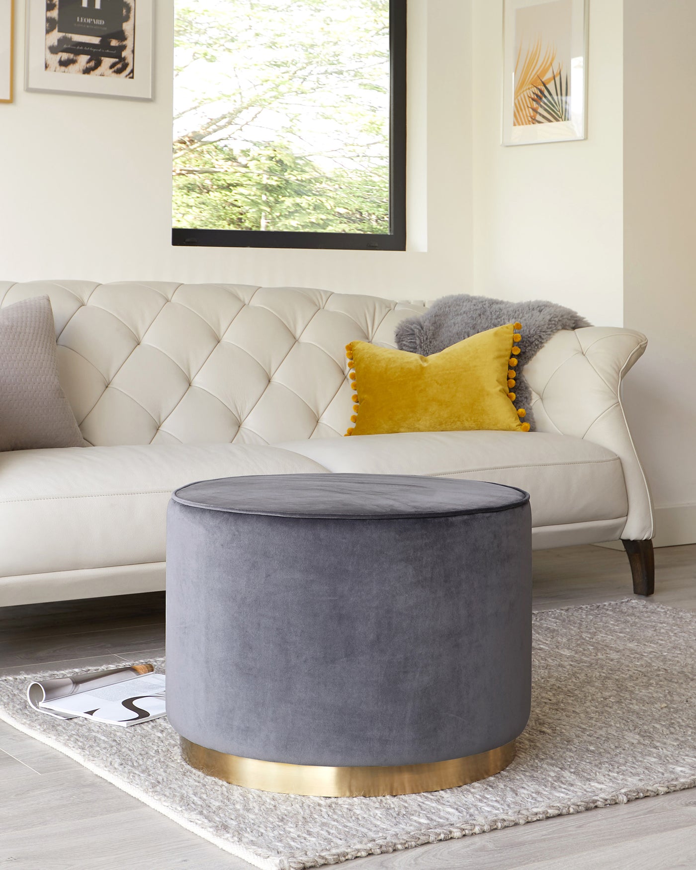 Elegant ivory tufted back sofa accompanied by a round, grey upholstered ottoman with a golden metal base, accented with colourful throw pillows and situated on a textured grey area rug.