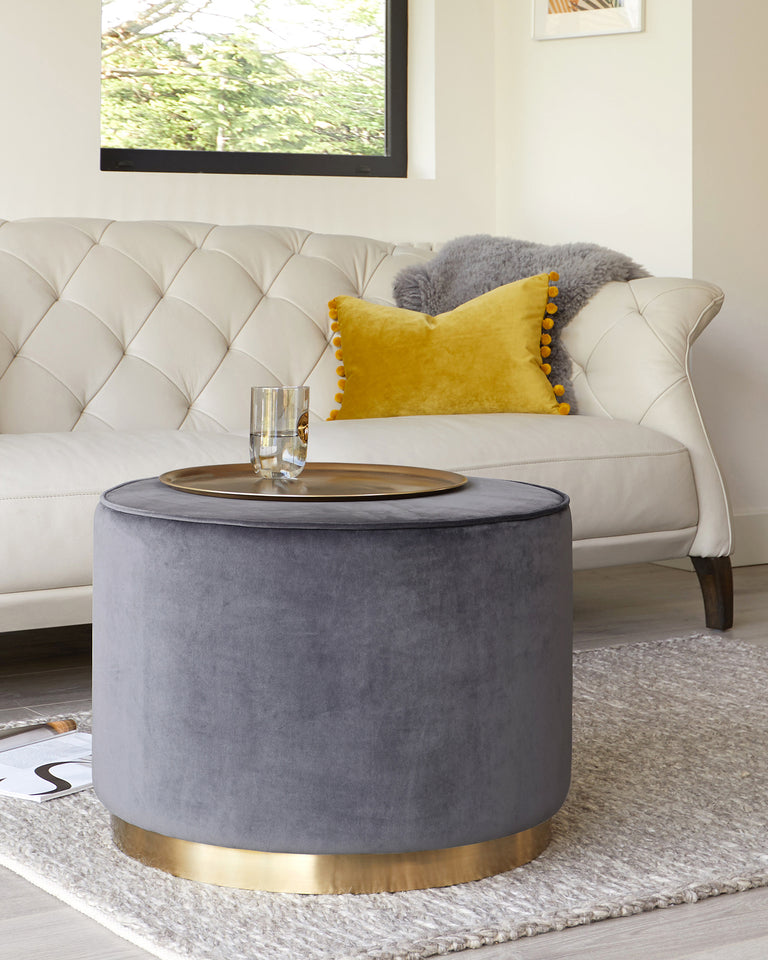 Elegant living room furniture featuring a tufted cream sofa with a curved back and dark wooden legs, complemented by a velvety grey round ottoman with a brass base. The ottoman is topped with a sleek round tray holding a clear glass. Two decorative pillows, one mustard with pom-pom edging and another grey faux-fur, add a touch of colour and texture to the sofa, all set against a neutral-toned rug with a subtle sheen.