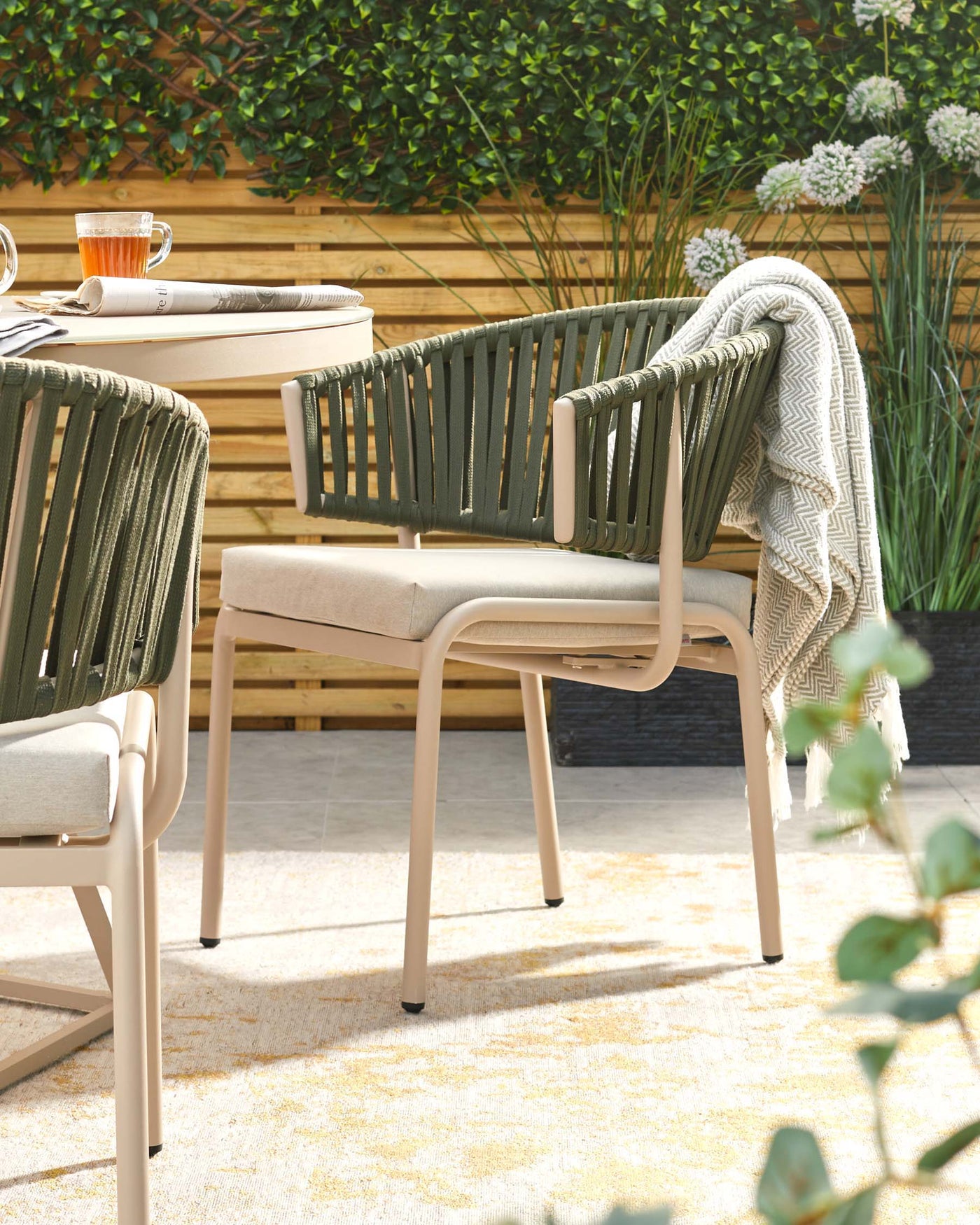 Modern outdoor furniture set featuring a beige metal frame chair with olive green rope backrest and armrests, complemented by a light beige seat cushion. A throw blanket is casually draped over one chair, signalling comfort. The round side table with a similar beige frame holds a glass cup and reading materials, creating a relaxing ambiance on a sunlit patio surrounded by lush greenery.