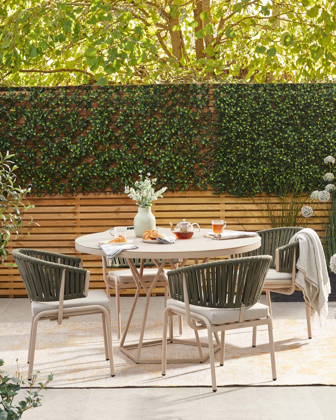Outdoor patio set with a round, beige table and four modern armchairs with olive-green straps and beige frames, complemented by light grey cushions. A beige and white patterned area rug lies beneath the set. The table is adorned with a vase of white flowers, teapot, cups, and pastries, creating an inviting scene for al fresco dining.