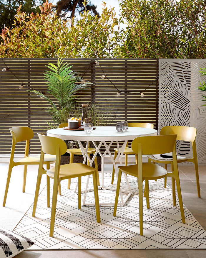 Round white outdoor dining table with a decorative leg design and four mustard yellow, modern-style chairs on a geometric-patterned outdoor rug.