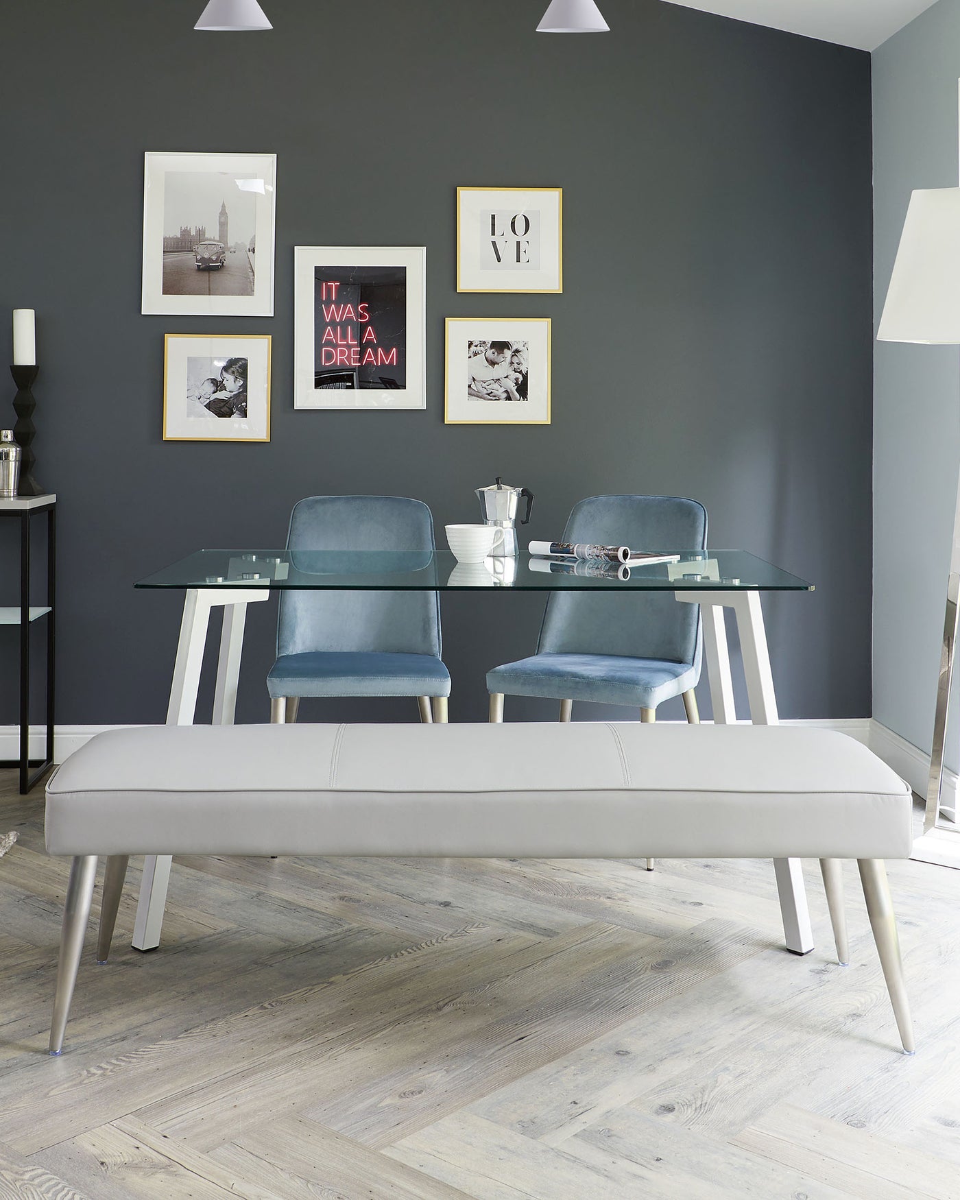 Modern dining room with a sleek glass-top table and four translucent blue dining chairs. A light grey upholstered bench with clean lines and subtle stitching details provides additional seating, all resting on a herringbone patterned wooden floor.