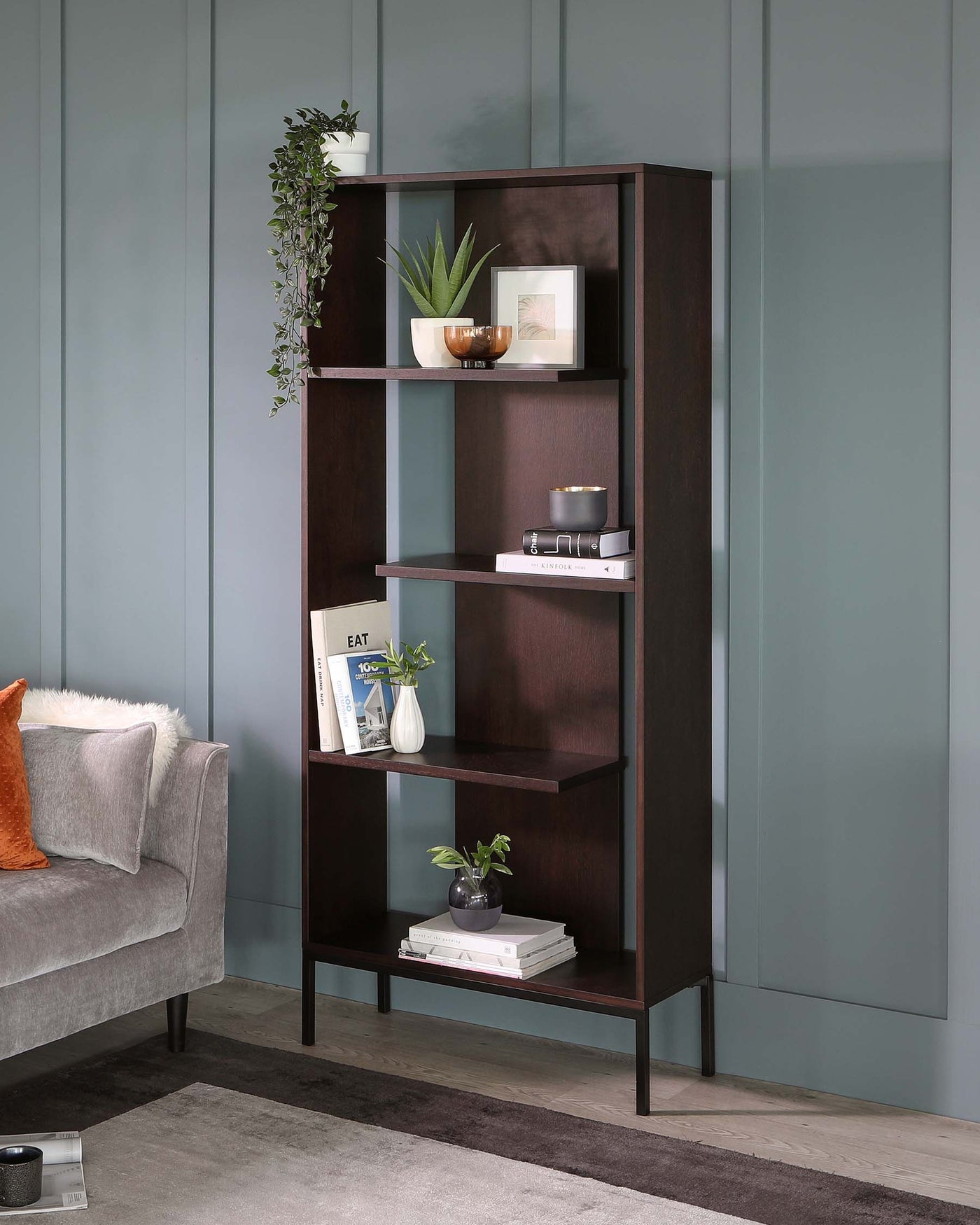 A modern five-tiered dark wood bookshelf with an asymmetrical design, featuring various decorative items on its shelves, against a teal wall. The shelving unit has a sleek silhouette with open back and sides, allowing for versatile placement in a room.
