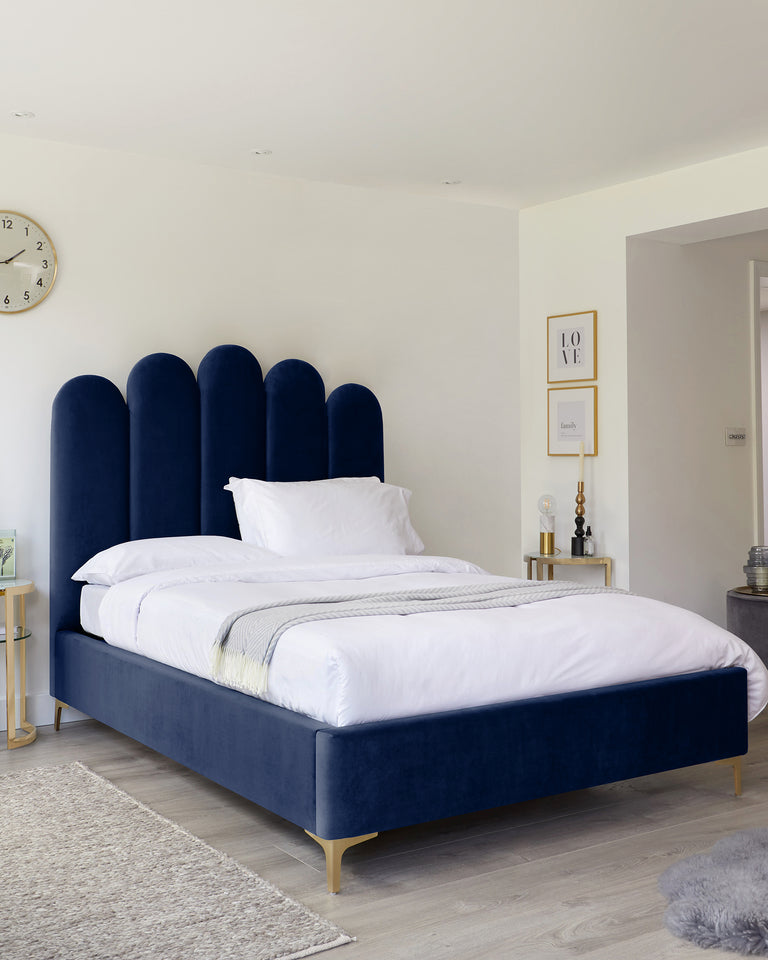 Elegant navy blue upholstered bed with a tall, unique rounded headboard and slender golden legs, accompanied by a small glass-top side table with golden accents. There is also a grey rug partially visible at the foot of the bed.