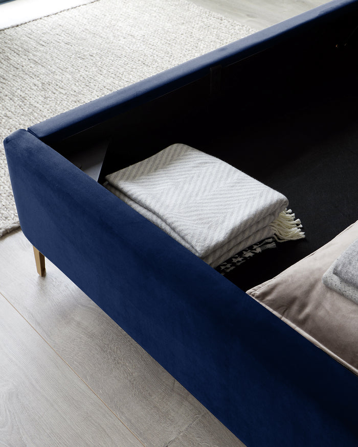 A modern navy blue storage ottoman with an open lid revealing neatly folded blankets inside. The ottoman features elegant brass-toned legs, and it sits on a pale wood floor with a textured off-white rug partially visible in the background.