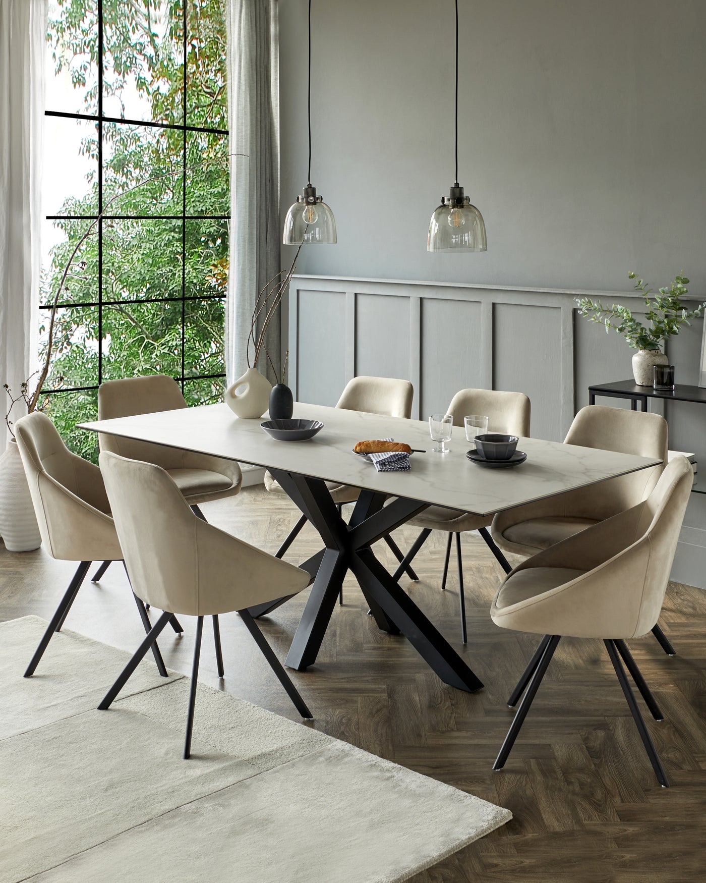 Modern dining room with a rectangular marble-topped table featuring a unique black crisscross base. Surrounded by six beige upholstered chairs with sleek black metal legs. A matching off-white area rug lies beneath the ensemble.