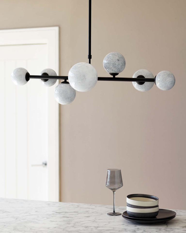 Elegant marble countertop with a set of simplistic, modern dishes in grey and black tones, paired with a smoky grey wine glass. Above, a contemporary linear chandelier features a series of unique, marble-patterned glass globes on a matte black horizontal bar, providing a minimalist yet chic overhead lighting solution.