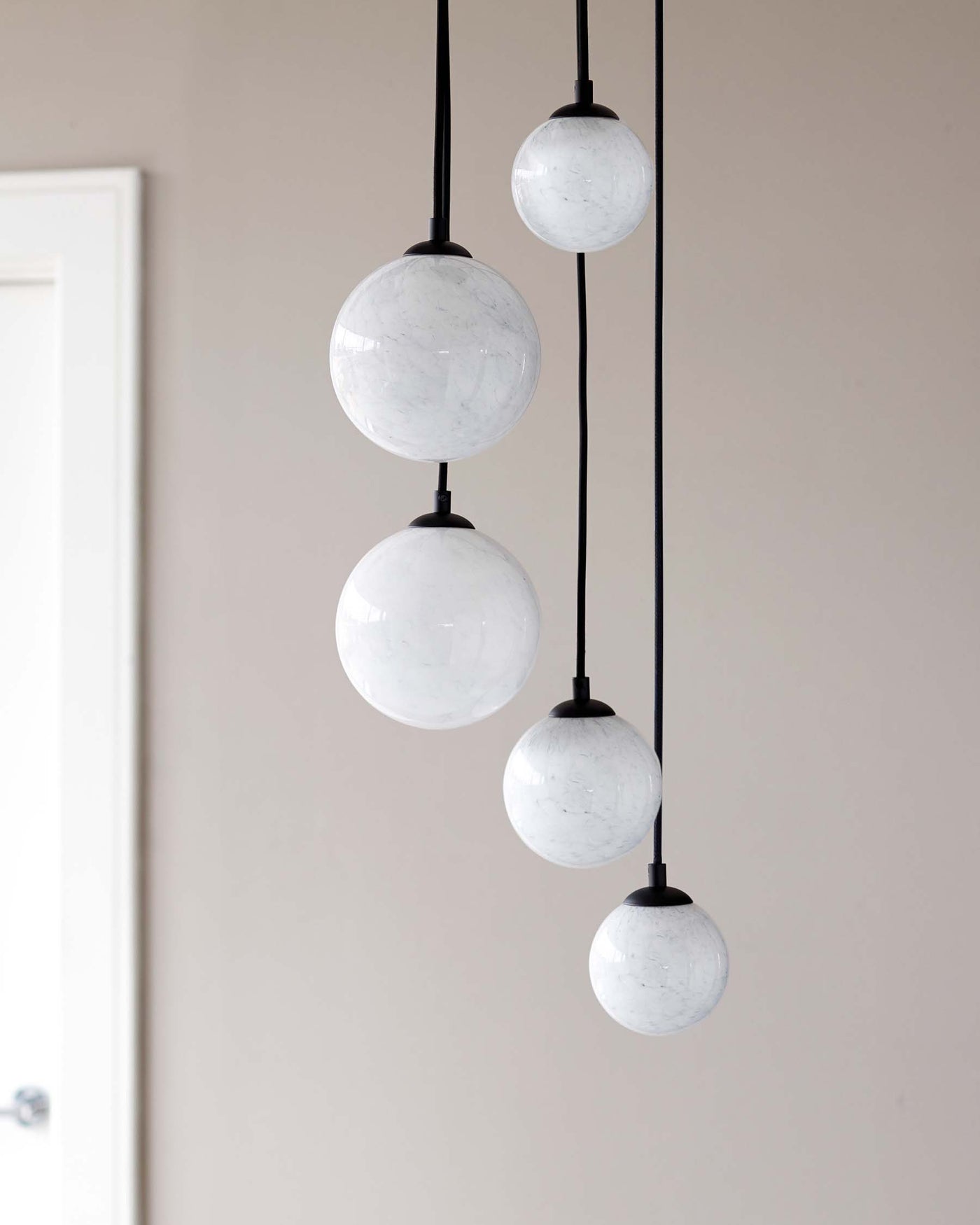Modern pendant light fixture with five spherical, white marbled glass shades of different sizes, suspended at varying heights by black cords against a light neutral background.